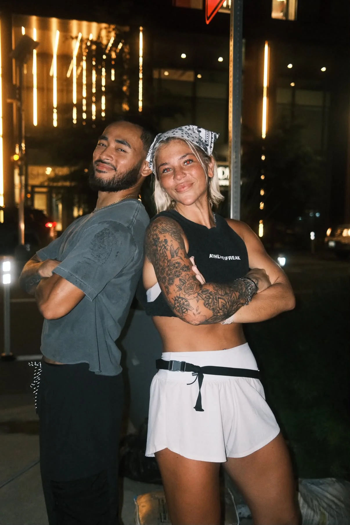 A man and a woman standing back to back with crossed arms, smiling at the camera outdoors at night, with city lights and a building illuminated in the background.