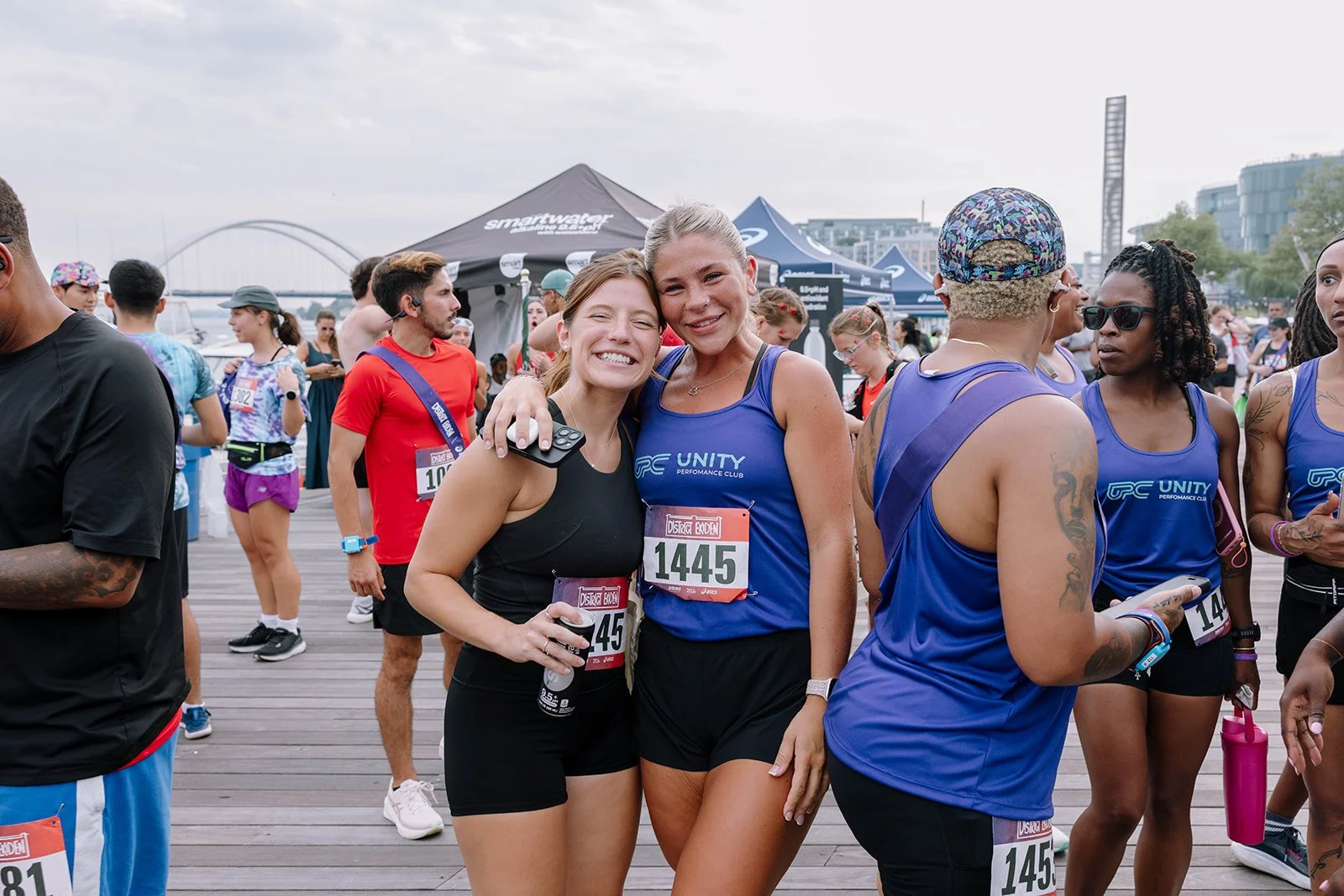 Two women smiling and hugging after a race, surrounded by other runners and spectators at an outdoor event in a city with modern buildings and tents.