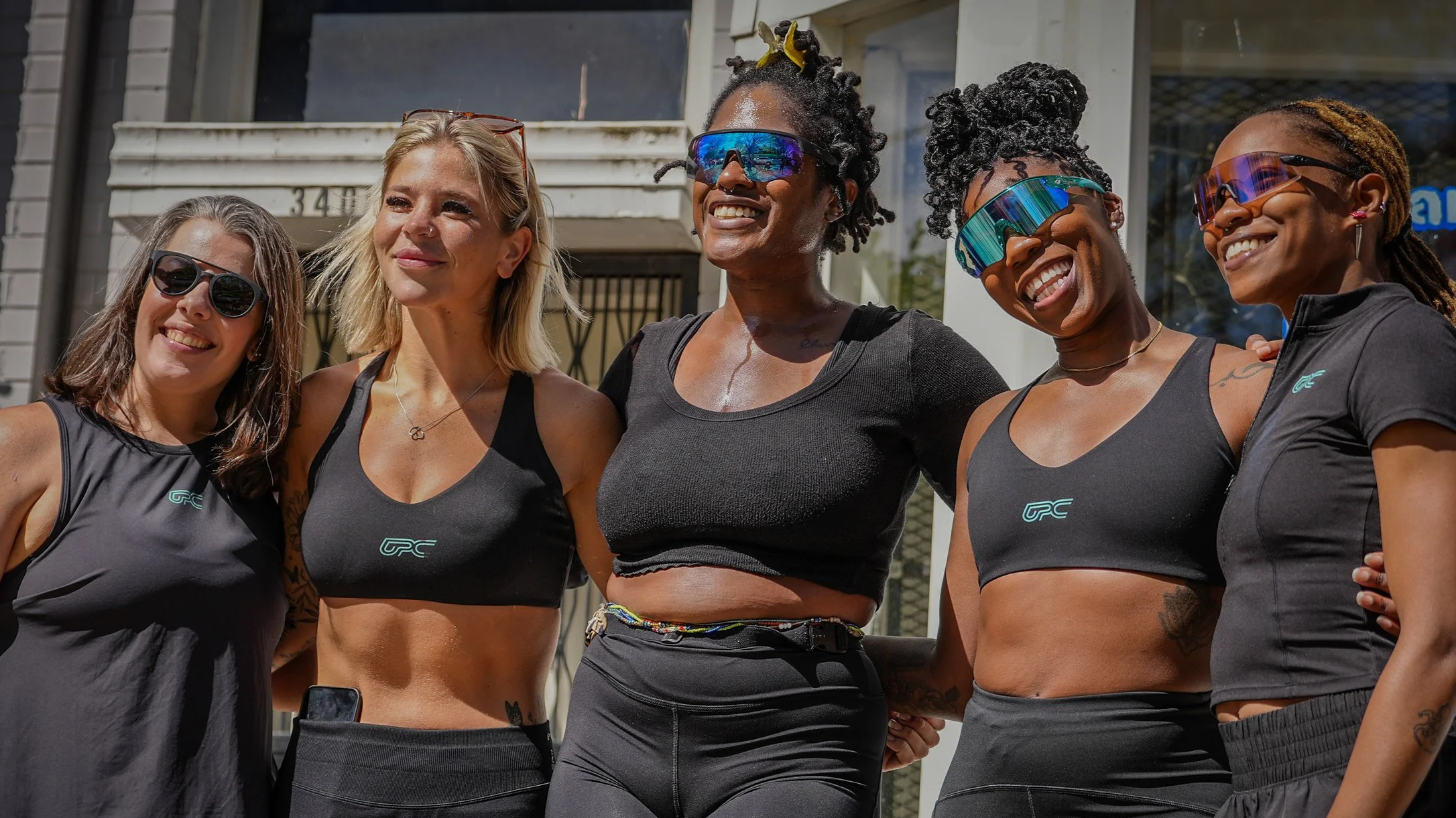 Group of five women wearing fitness attire and sunglasses standing outdoors, smiling and posing for a photo.