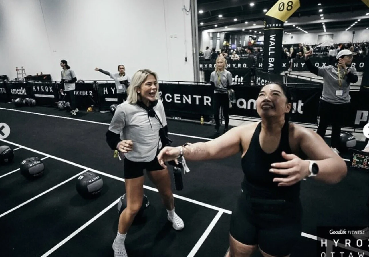 Two women in a gym, one smiling with her hand on her chest and the other holding her hand in a shake, surrounded by gym equipment and other women in workout attire, with a busy fitness event in the background.