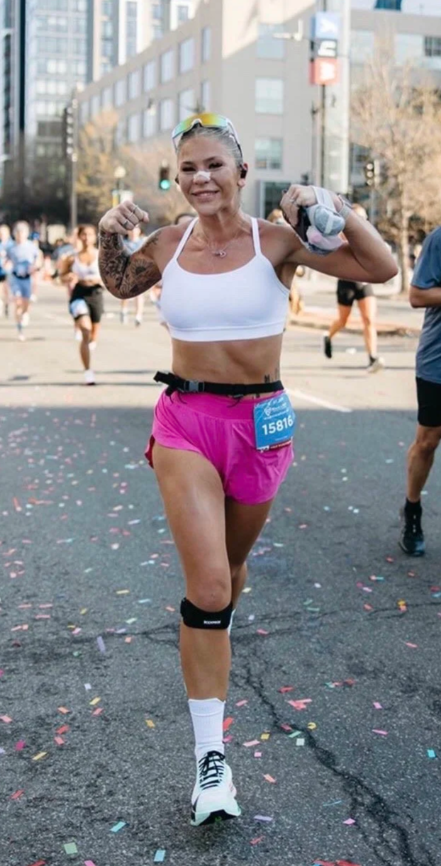 A woman running in a marathon, flexing her arms and smiling, wearing a white sports bra, pink shorts, and a knee brace, with other runners and city buildings in the background, and colorful confetti on the ground.