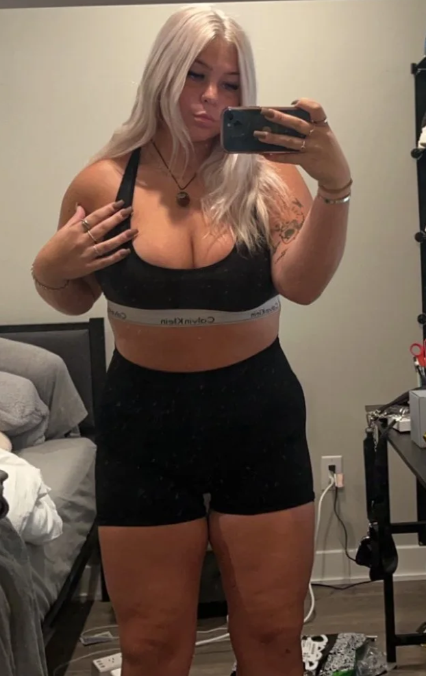 A woman with long blonde hair taking a mirror selfie in a bedroom, wearing a black sports bra and black shorts.