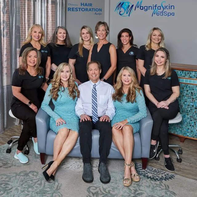 Group photo of eleven people, predominantly women, inside a dental clinic, with the sign 'My Magnifa Skin & Med Spa' and 'Hair Restoration' visible in the background. Some are seated on a sofa, others stand behind, all smiling.