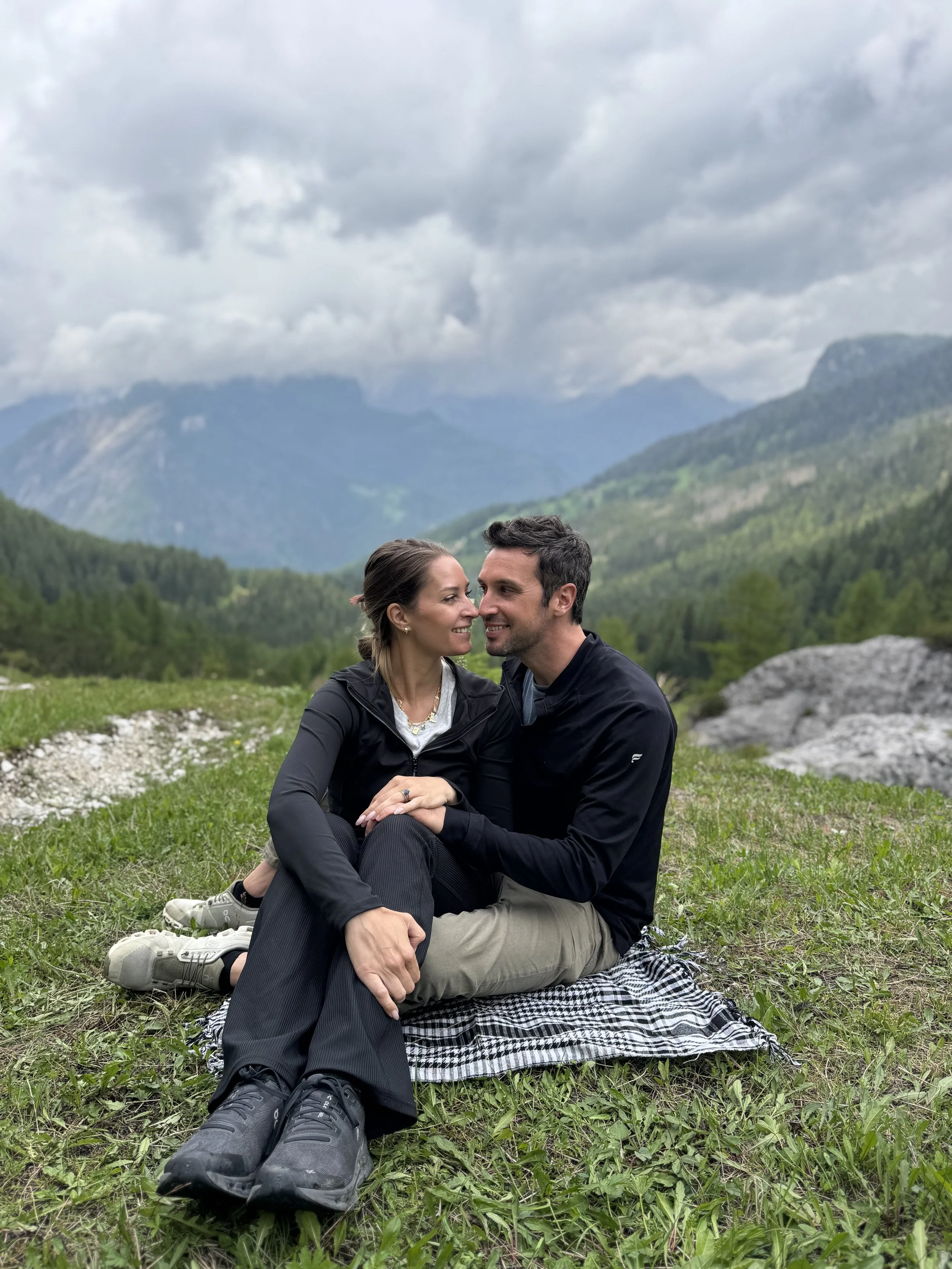 Couple looking at each other, sitting on green grass with blue sky and mountains in the background