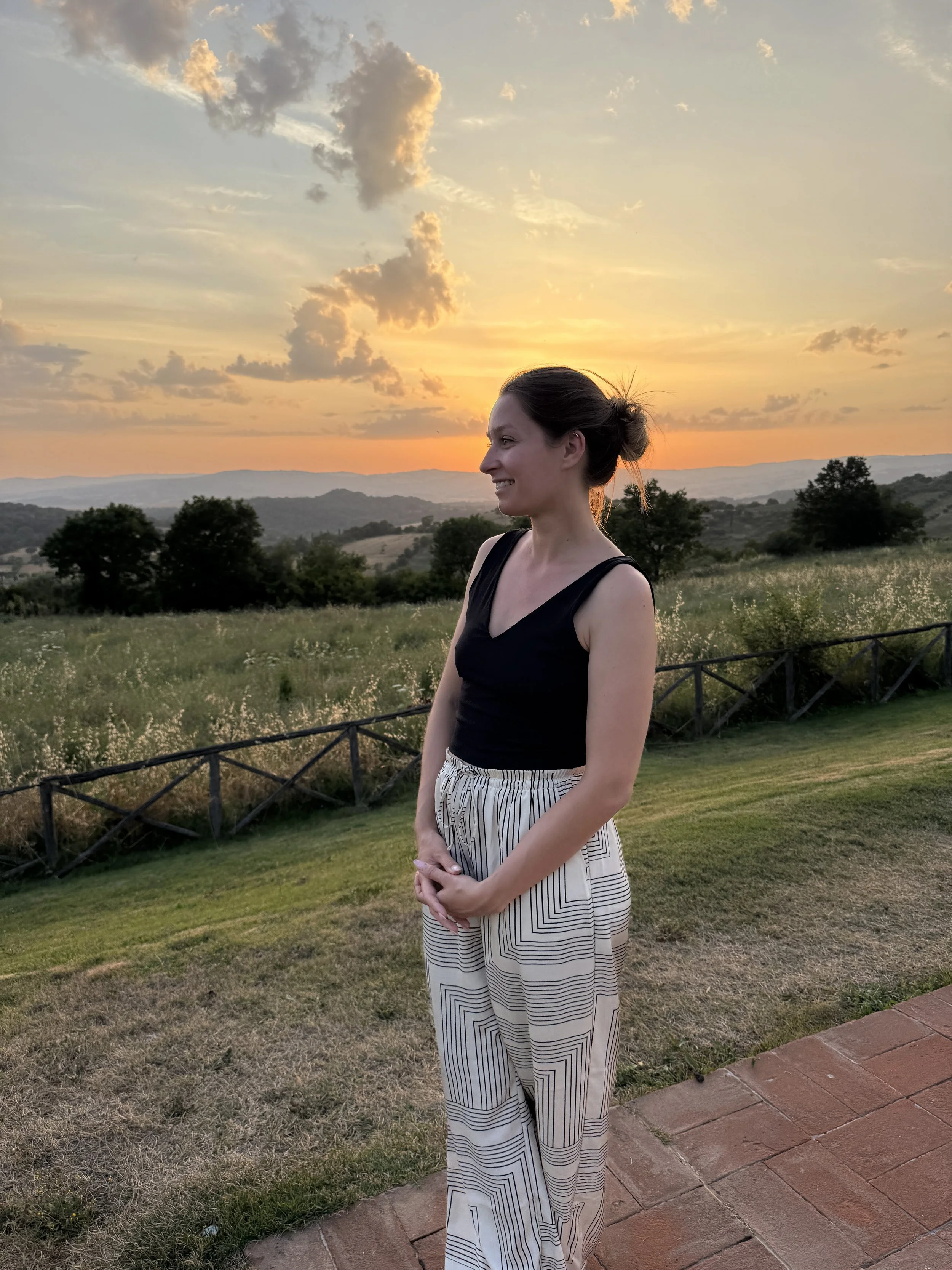 Womean in black and white clothes looking out over a vineyard in italy with the sunsetting behind her