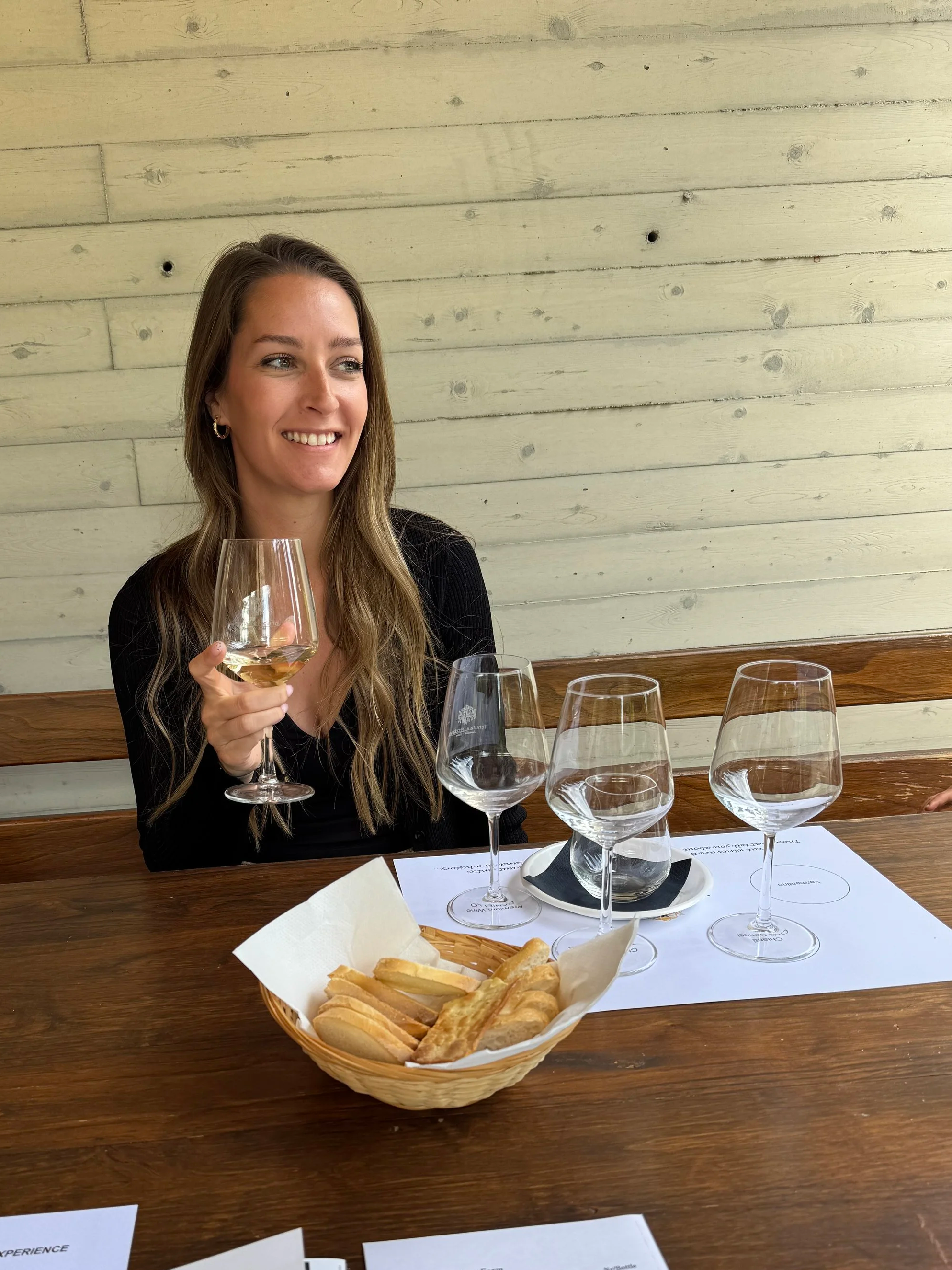 A woman sitting at a wooden table, holding a glass of white wine, with a basket of bread and several empty wine glasses in front of her. The background features a light-colored wooden wall.