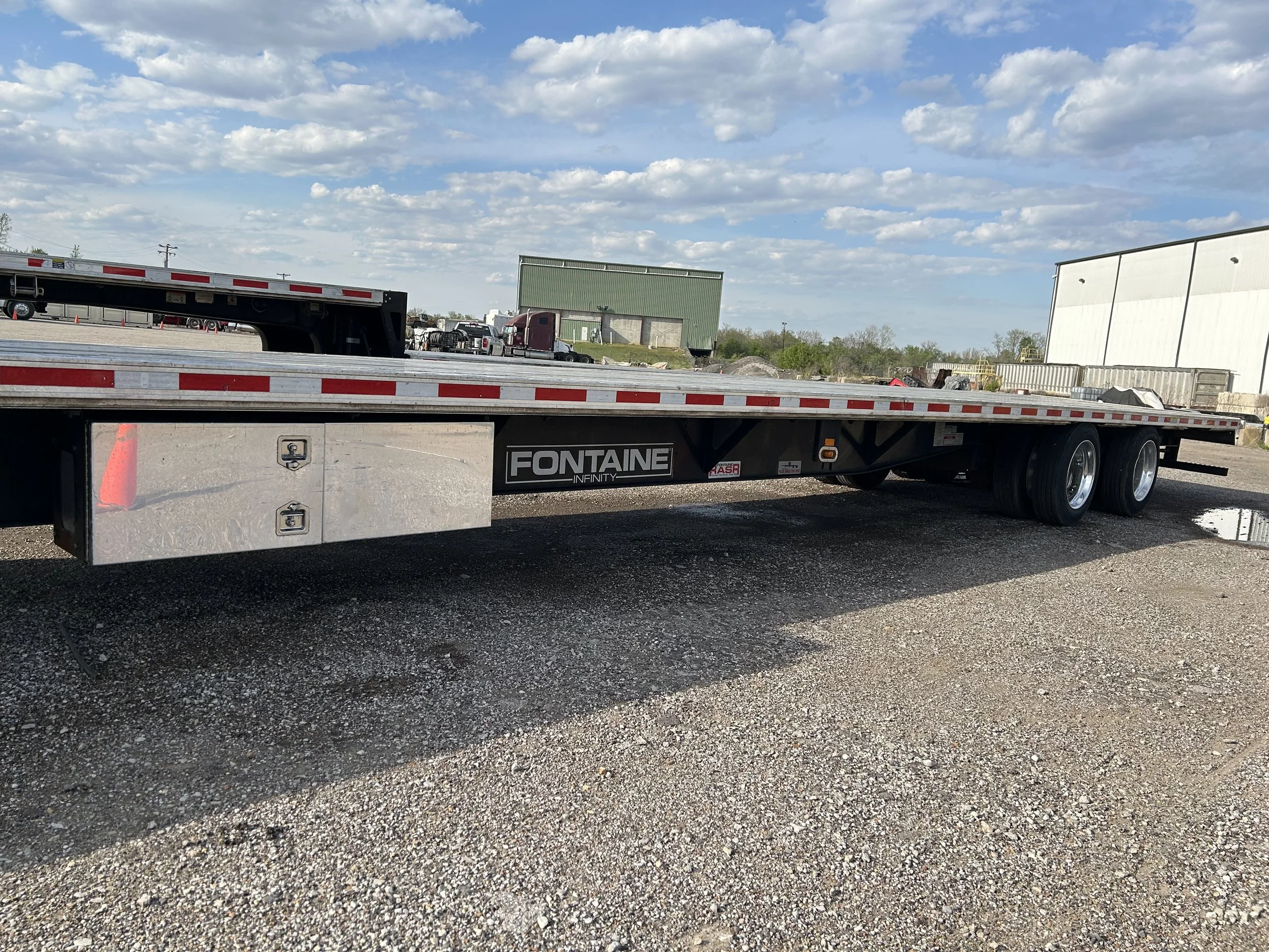 A flatbed trailer with the brand name Fontaine parked on gravel, with a partly cloudy sky and industrial buildings in the background.