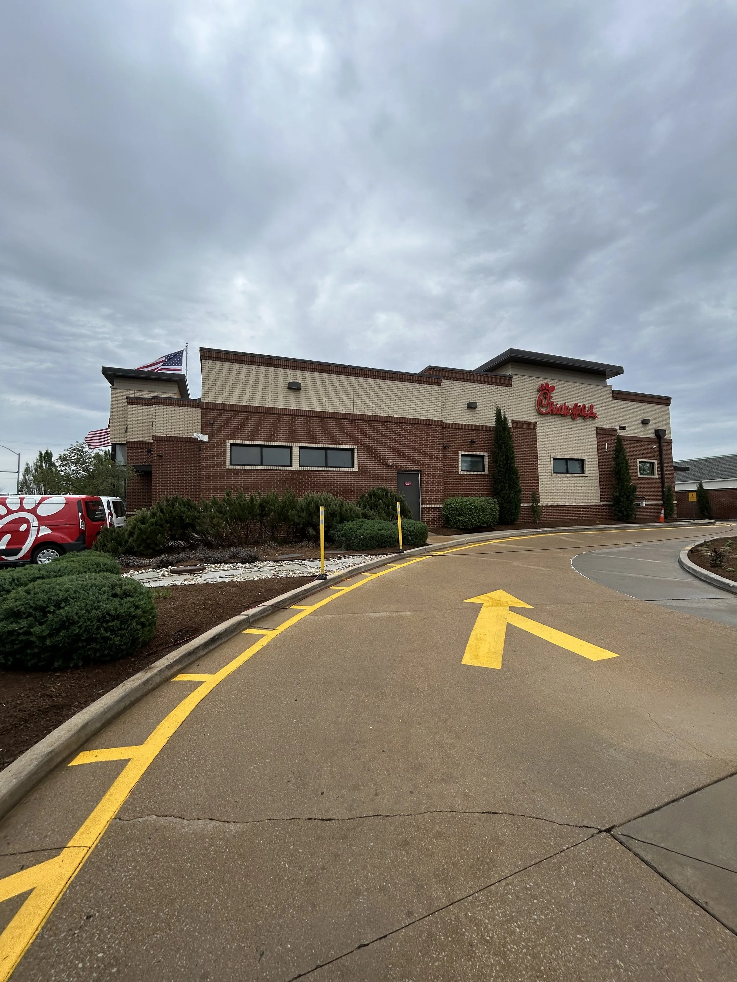 View of a Chick-fil-A restaurant building with American flags, a parking lot, and a yellow directional arrow on the pavement, cloudy sky overhead.