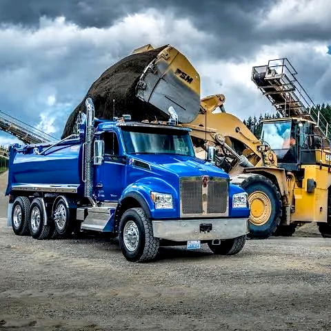 A large blue dump truck and a yellow bulldozer working together outdoors during daytime with cloudy skies.