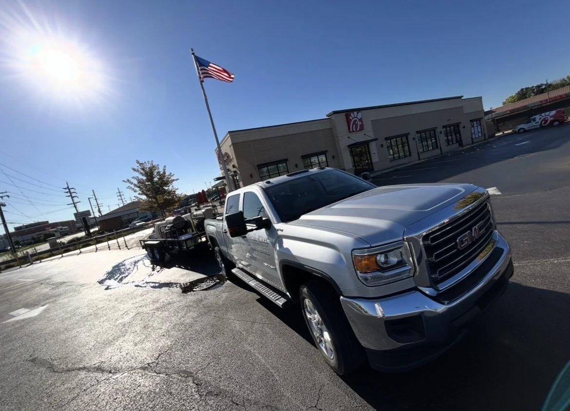 A silver GMC pickup truck with a trailer attached, parked in a parking lot near a Chick-fil-A restaurant, under a sunny sky with an American flag flying.