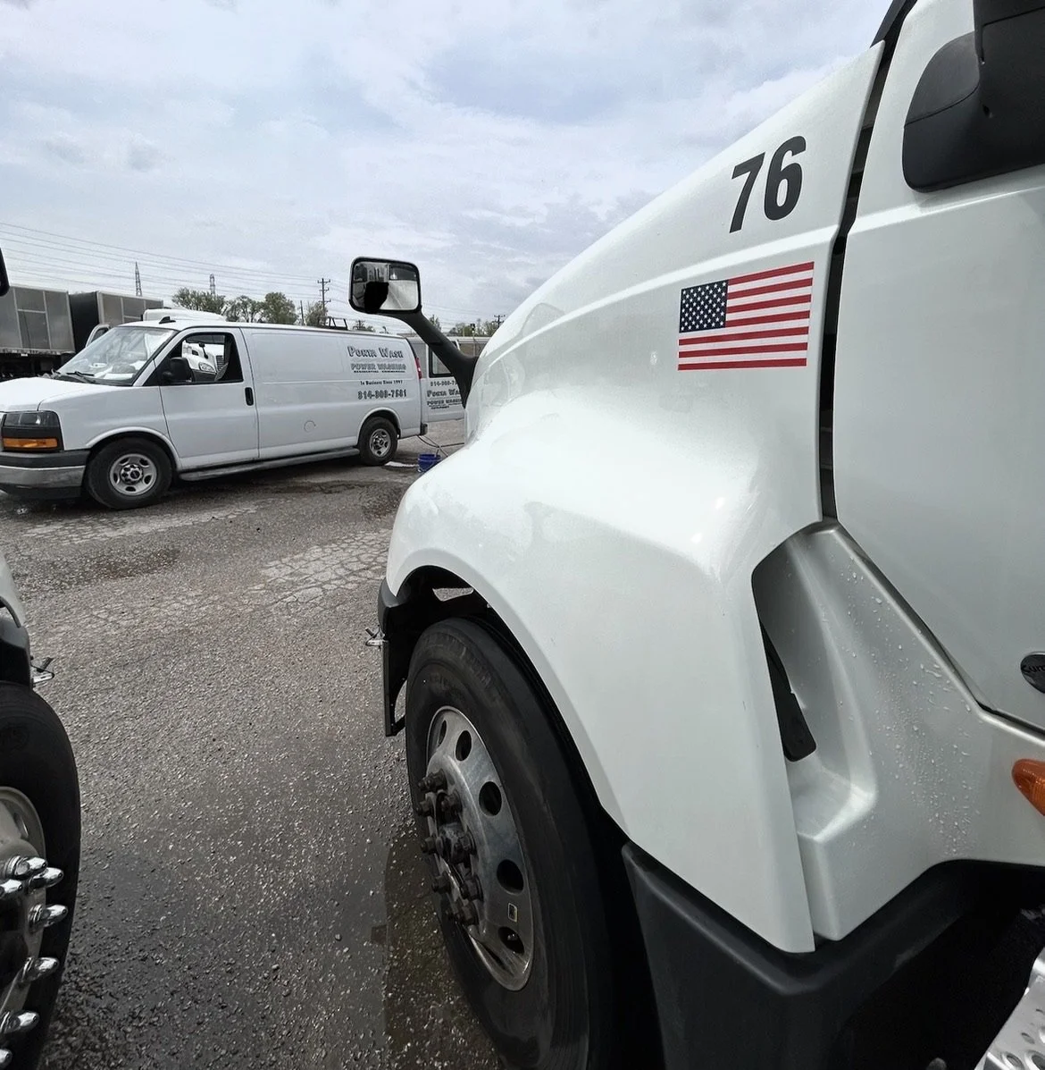 A parking lot with trucks and vans, including a white semi-truck with an American flag decal and the number 76, and a smaller white van with business information on its side.