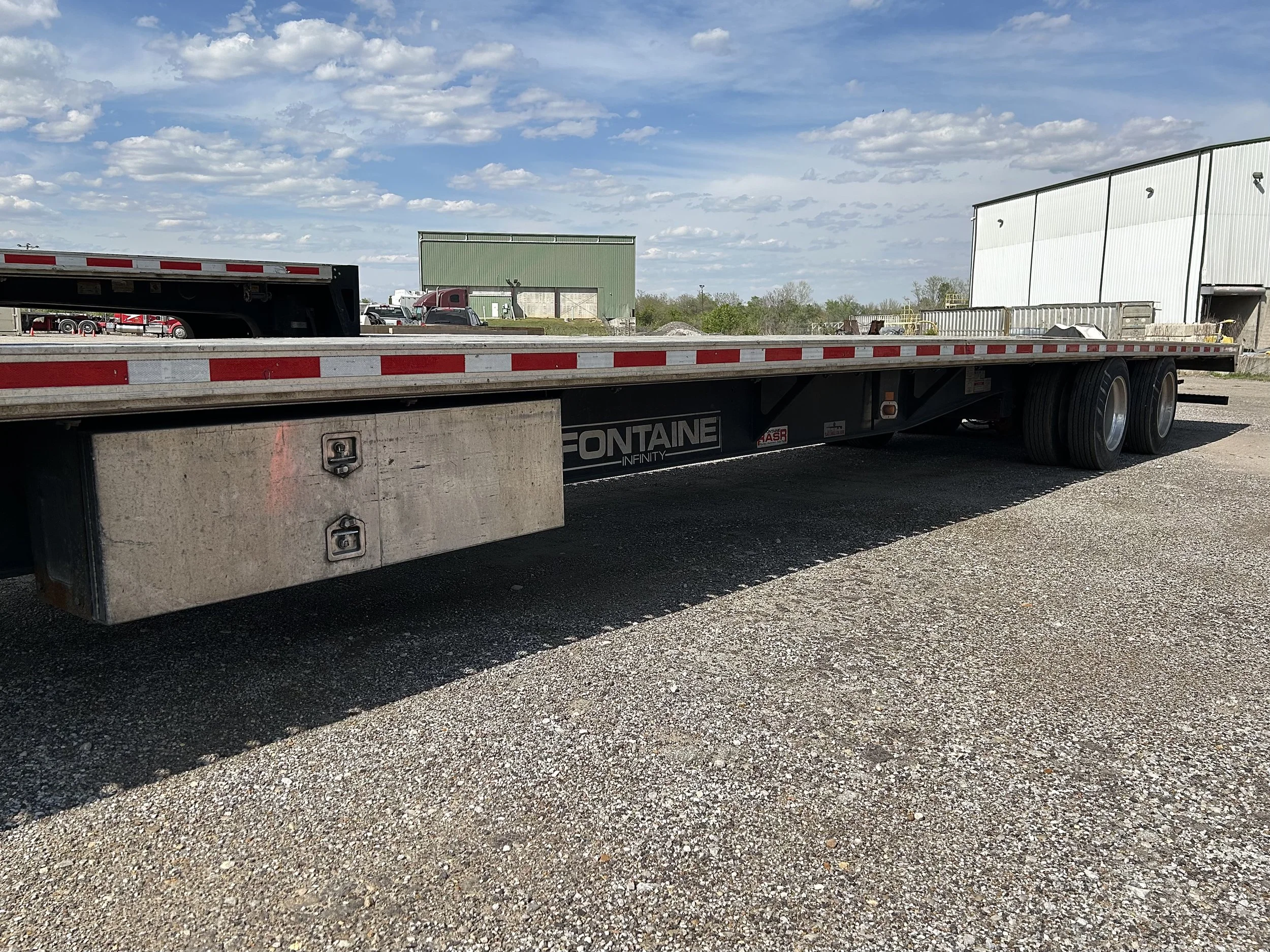 Flatbed truck trailer parked on gravel ground with a clear sky and industrial buildings in the background.