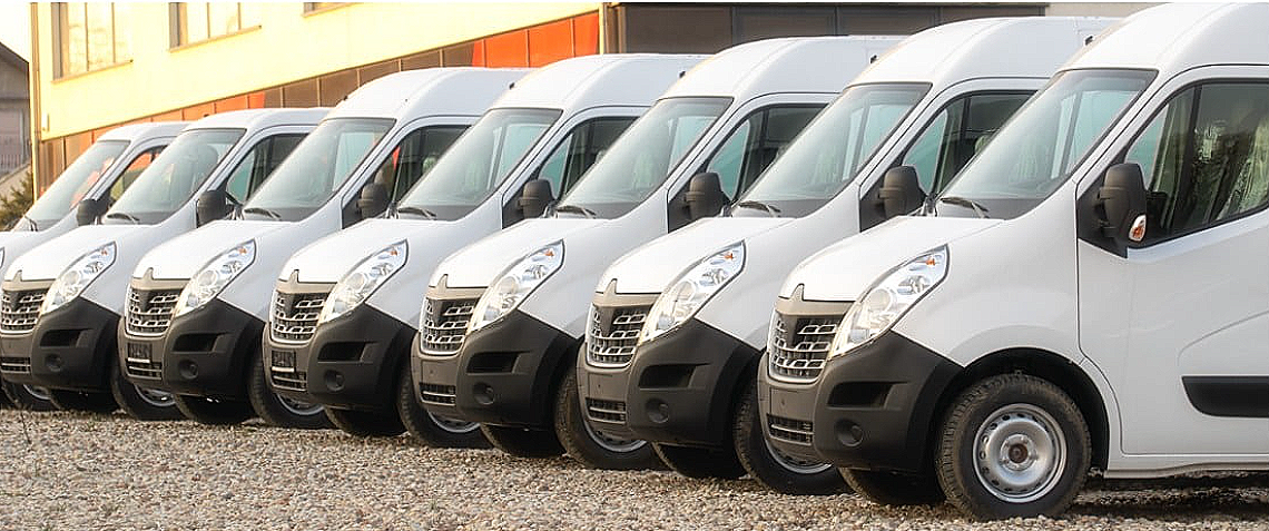 A row of white cargo vans parked on a gravel lot outside a building.