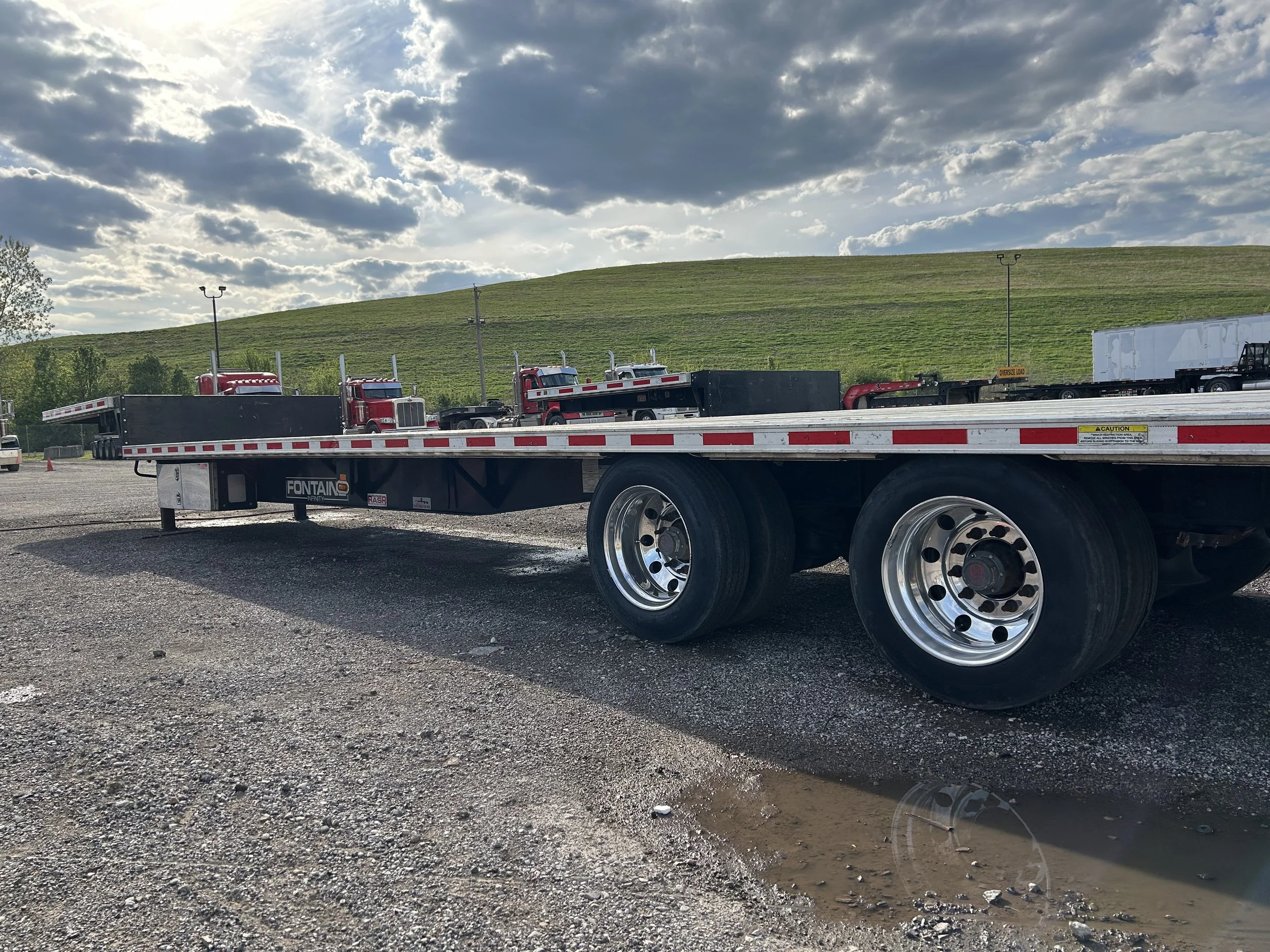 A flatbed truck trailer parked on gravel ground with other trucks in the background and a grassy hill under a cloudy sky.
