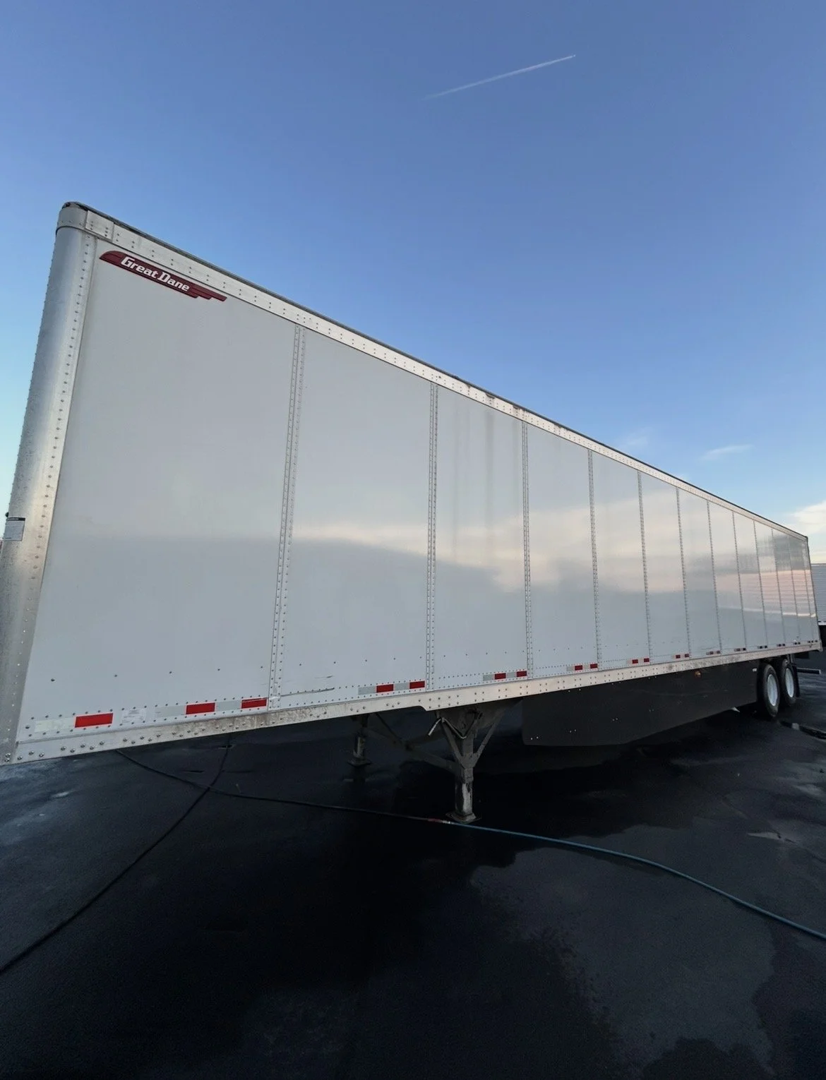 Large white semi-trailer truck parked on a paved surface under a clear blue sky with a contrail.