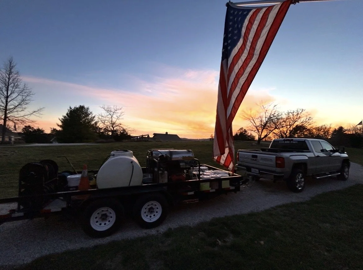 A pickup truck with a trailer attached, carrying barbecue equipment, is parked outdoors at sunset. An American flag is hanging from a pole, with a scenic sky in the background.
