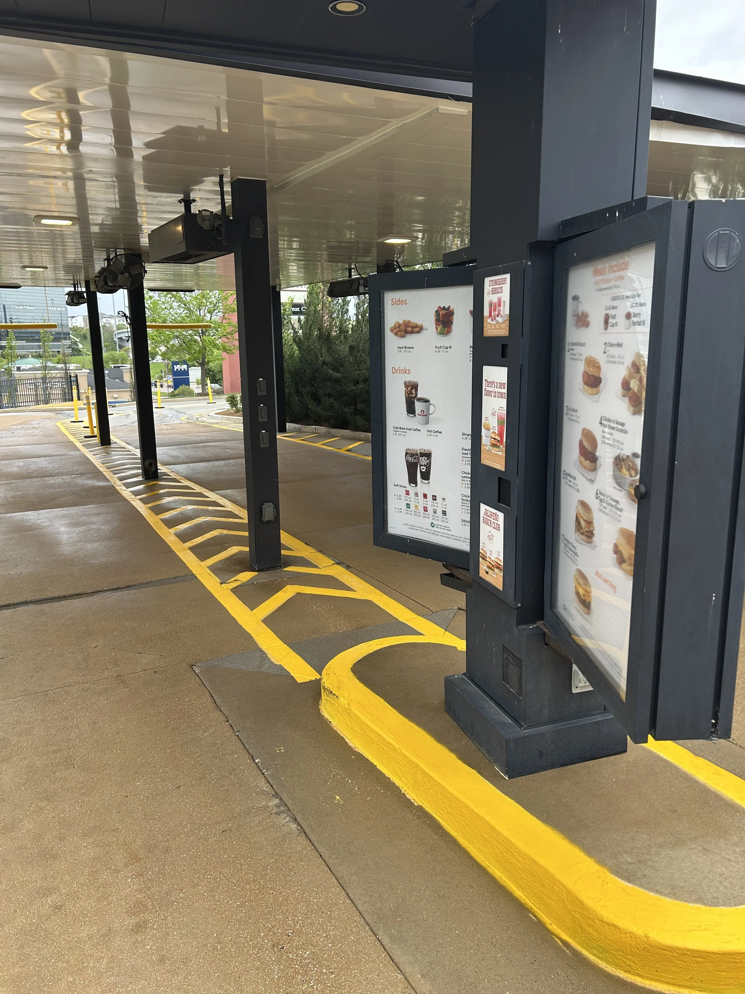 The image shows an empty drive-thru lane outside a fast food restaurant. There are two menu boards with food pictures and prices, black support poles, and yellow painted lines on the wet pavement denoting the drive-thru path. A building with a metallic roof and some trees can be seen in the background.