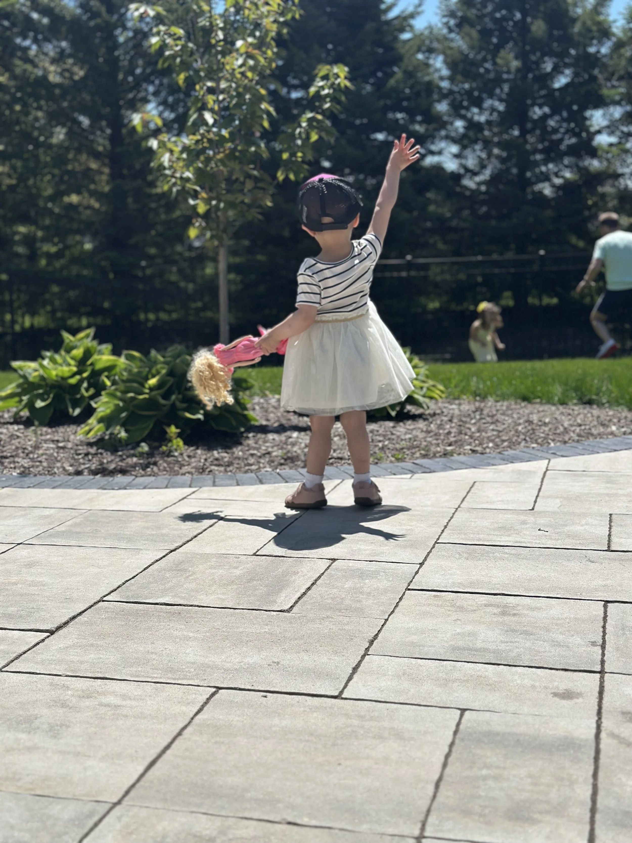 A young girl in a striped shirt, white skirt, and a black cap stands on a paved walkway, raising her right arm and holding a pink stuffed animal in her left hand, outdoors with trees and grass in the background.