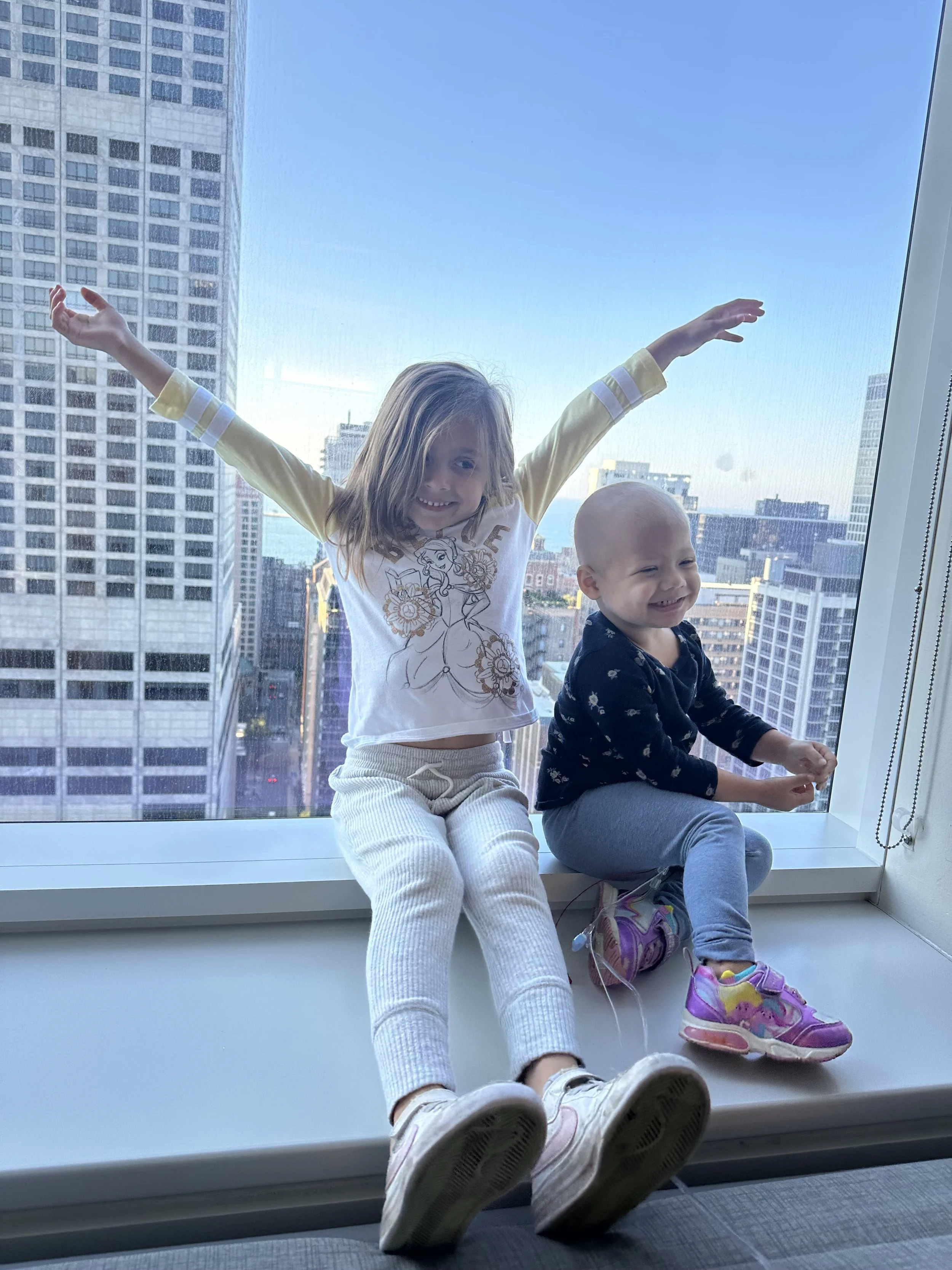 Two young children, a girl and a boy, sitting on a large windowsill with a cityscape of tall buildings behind them. The girl has her arms raised and is smiling, wearing a white shirt with a character drawing, cream pants, and white sneakers. The boy is sitting beside her, also smiling, wearing a dark patterned shirt, light gray pants, and purple shoes.