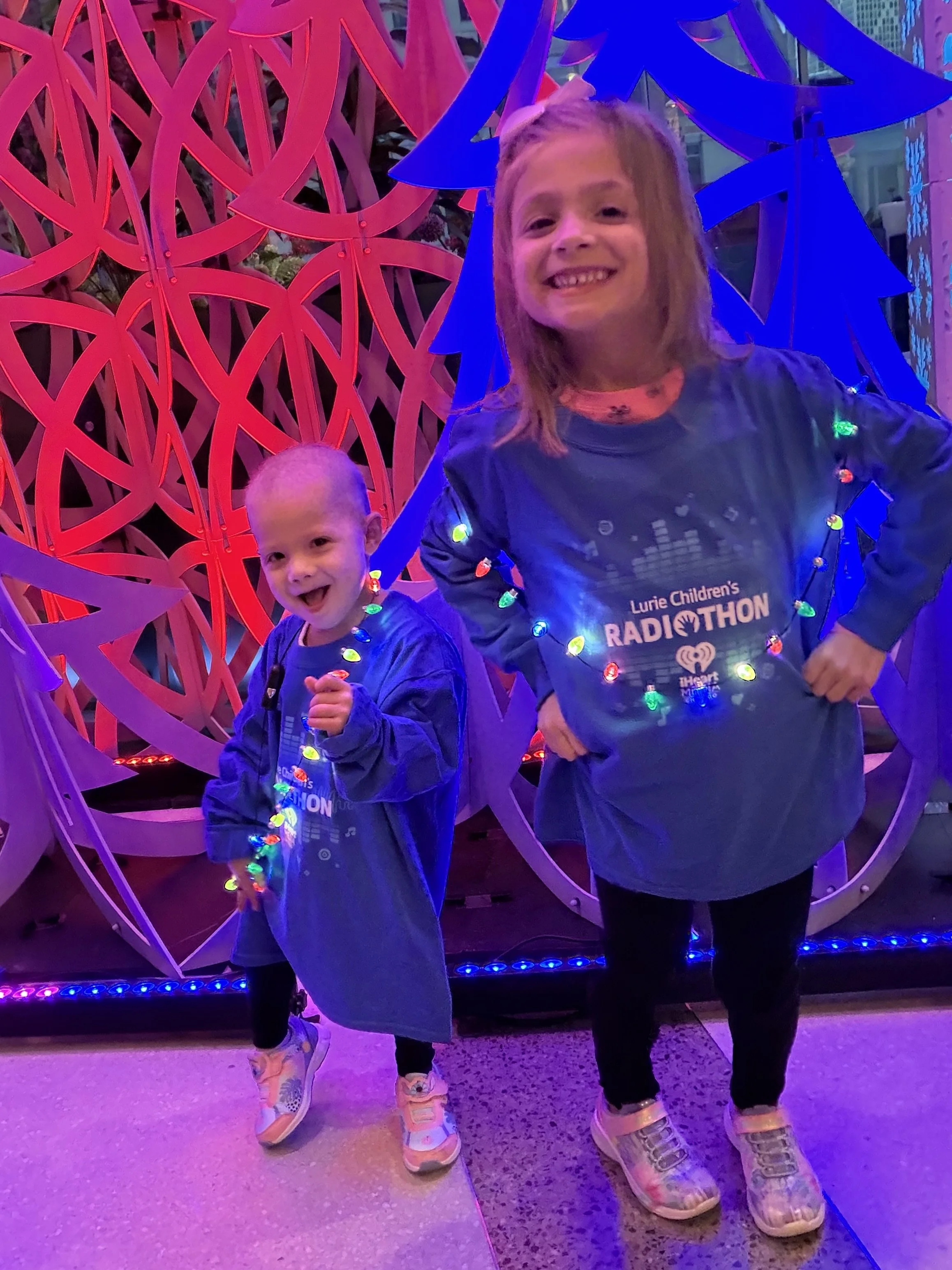 Two children smiling at a holiday event, wearing 'Lurie Children's Radiothon' shirts, surrounded by colorful holiday lights and decorations.