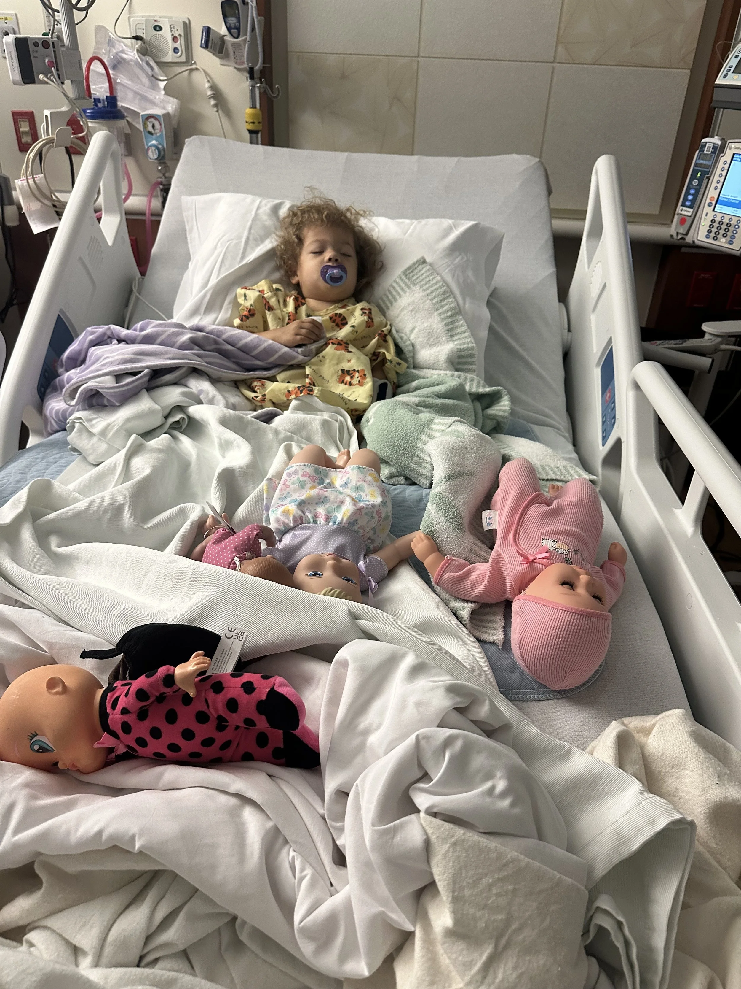 A young child with curly hair and a pacifier lying in a hospital bed surrounded by dolls and stuffed toys, with medical equipment in the background.
