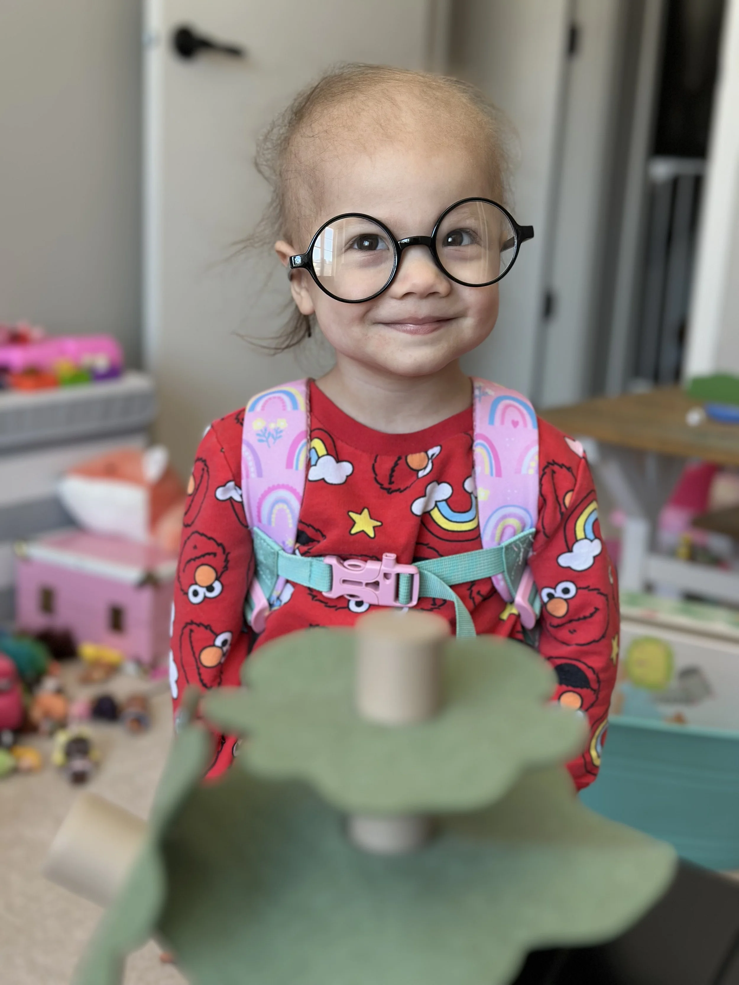 A young girl with glasses, wearing a rainbow and cloud-themed backpack and a Elmo-themed shirt, stands indoors with her cheeks slightly puffed out and a small smile, with toys and furniture in the background.