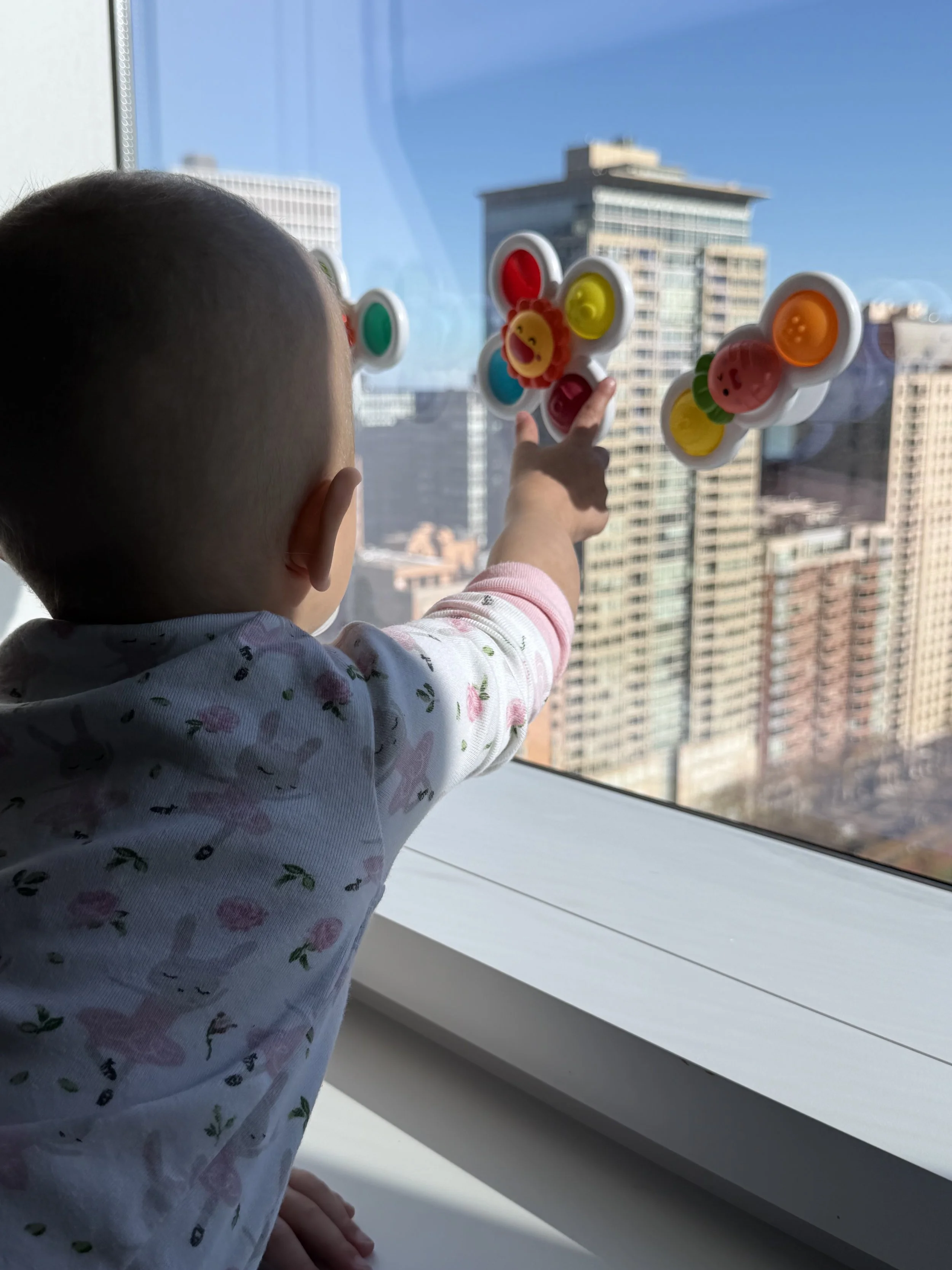 A young child reaching out with their hand, touching or pointing at a colorful fidget toy on a large window with a city skyline in the background.