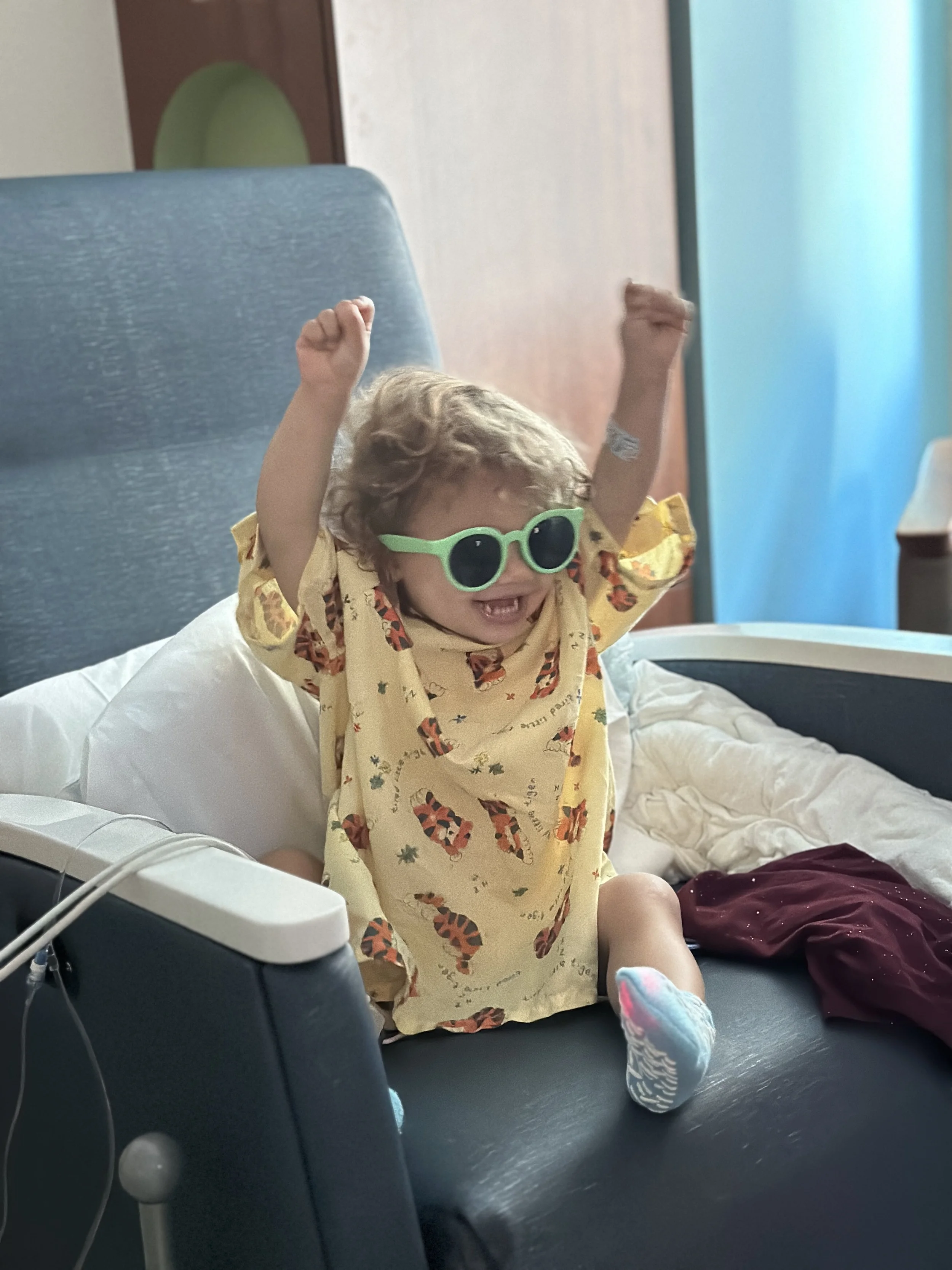 A young child with curly hair is sitting on a hospital bed wearing green sunglasses and a yellow shirt with animal prints. The child is smiling and raising their arms in excitement.