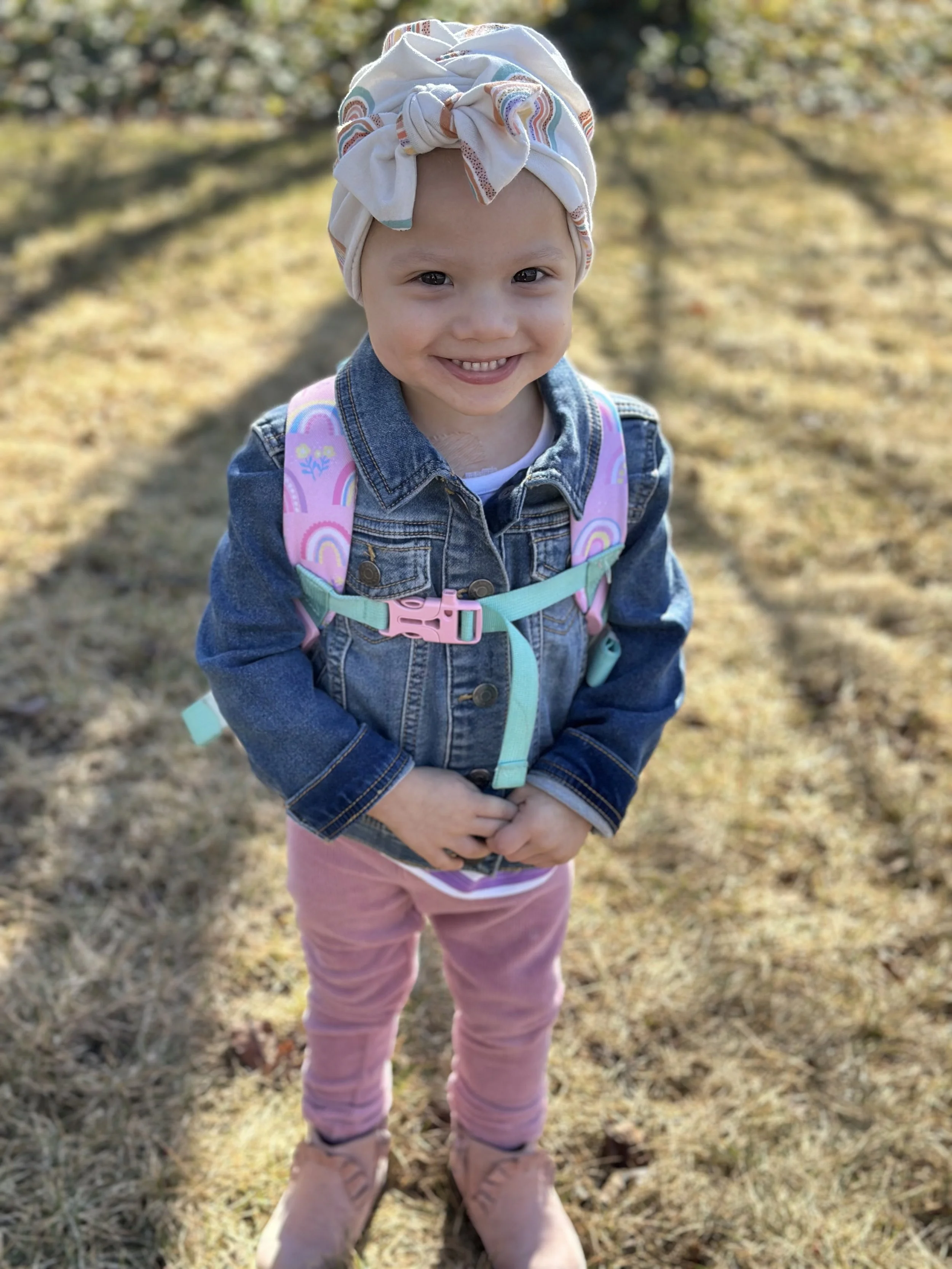 A young girl smiling outdoors, wearing a headscarf, denim jacket, pink pants, tan boots, and a pink backpack with rainbow designs.