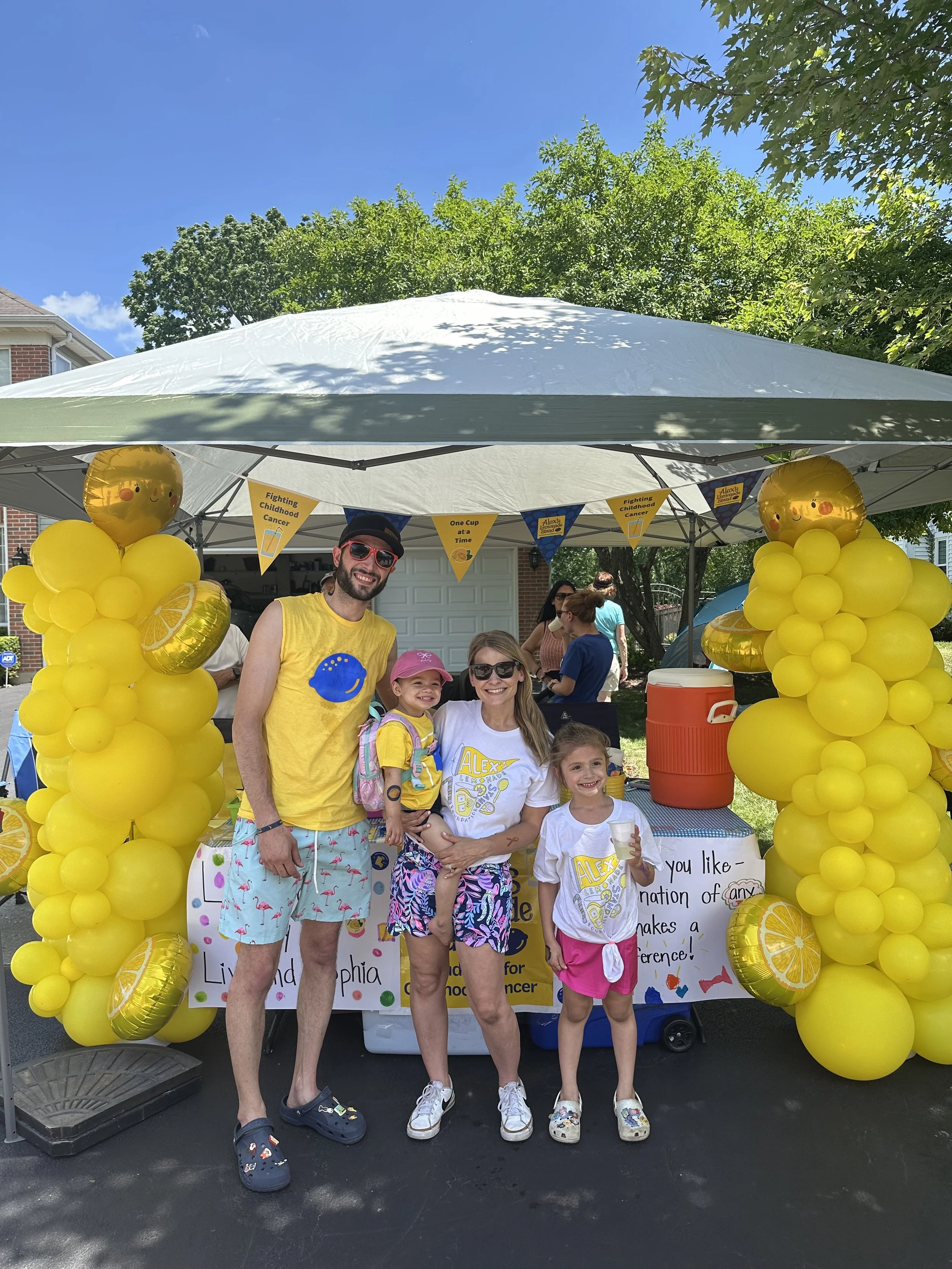 Family standing in front of a booth decorated with yellow balloons and banners promoting childhood cancer awareness at an outdoor event on a sunny day.