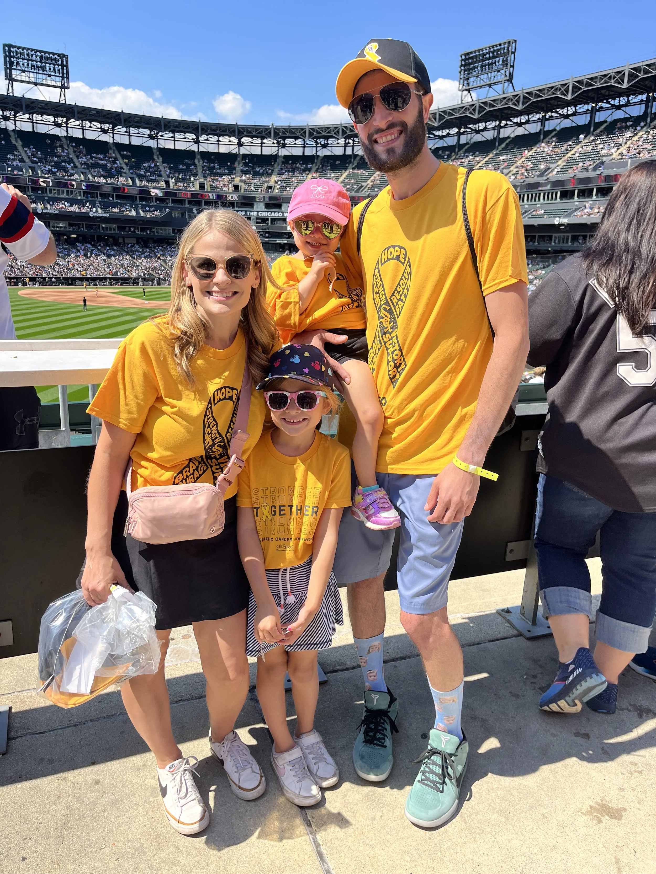 A family of four at a baseball game, wearing yellow shirts with a hope ribbon, standing on the stadium's sidewalk with the baseball field in the background. The father is tall with sunglasses, the mother is smiling, and two daughters wearing sunglasses are with them. The older daughter is standing and the younger daughter is sitting on her father's arm, all enjoying a sunny day at the stadium.