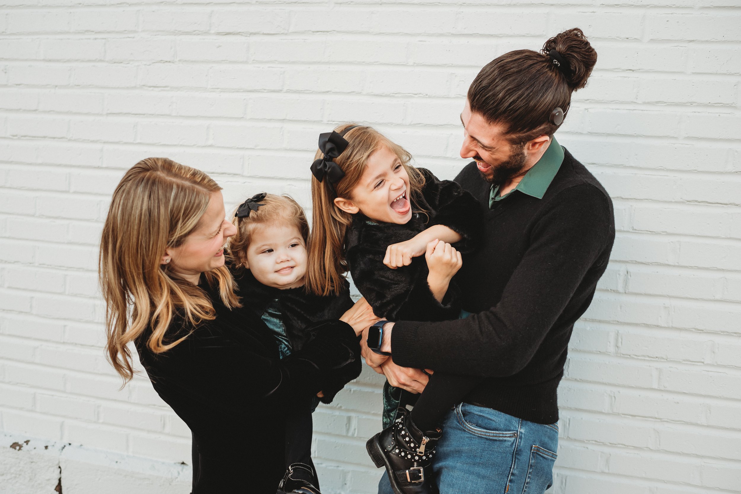 A man and a woman laughing and playing with three young girls in front of a white brick wall.