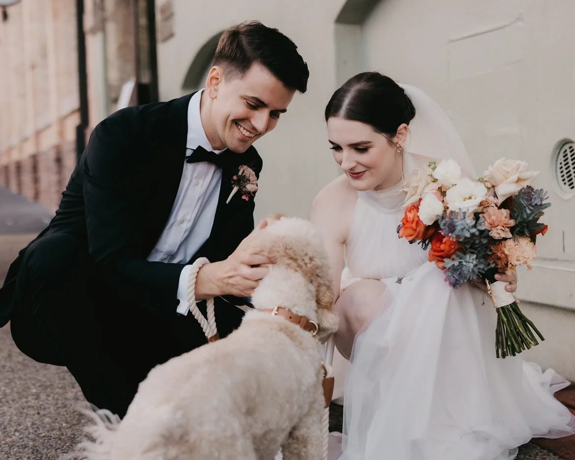 A bride and groom in wedding attire kneel outdoors, smiling at a golden retriever puppy between them. The bride holds a colorful bouquet, and the groom has a boutonniere, both appear joyful.