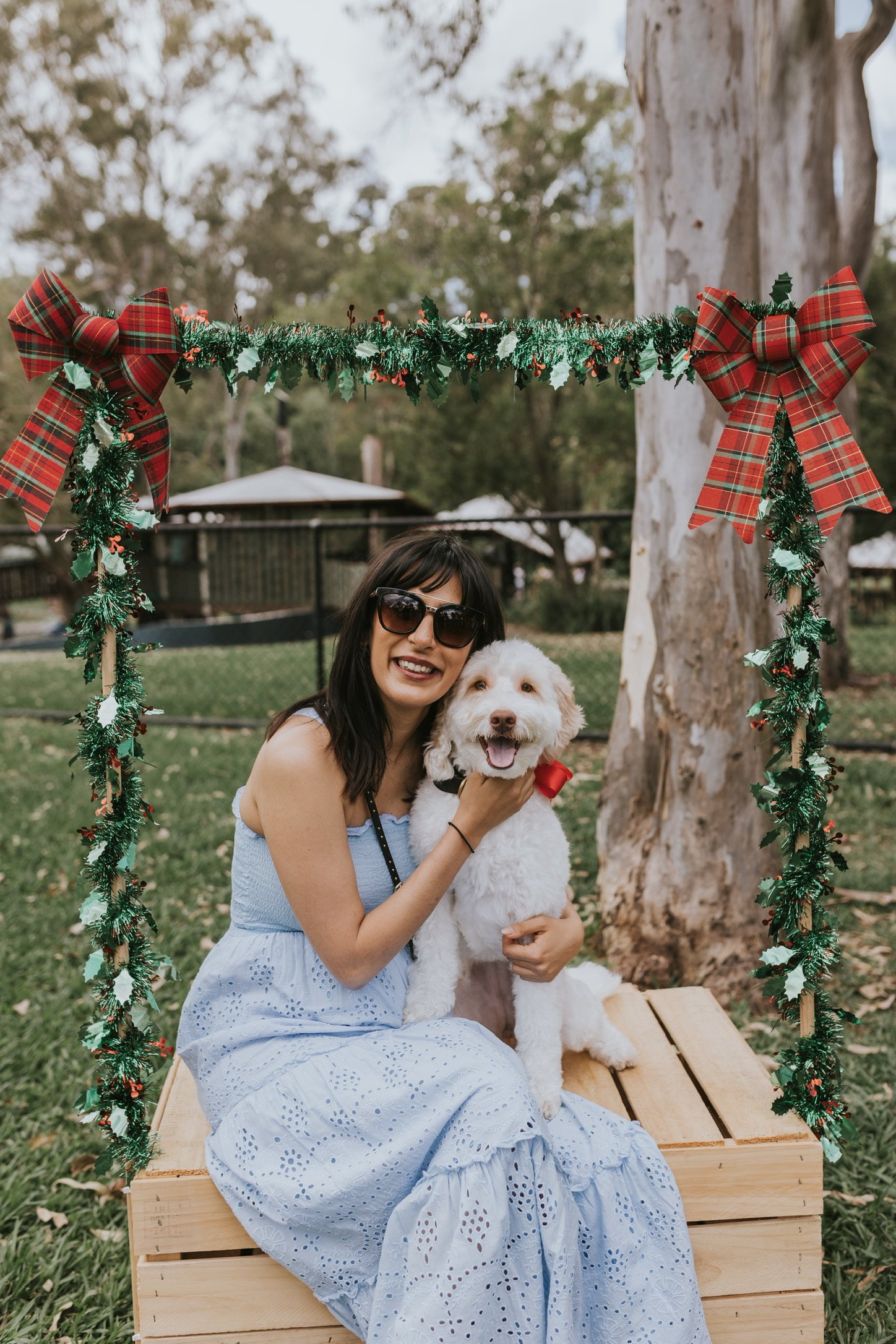 A woman with dark hair and sunglasses, wearing a light blue dress with eyelet embroidery, sitting on a wooden bench outdoors, holding a happy, white, fluffy dog with a red bow, under a decorated arch with green leaves and red plaid bows, in a park with trees and a fence in the background.