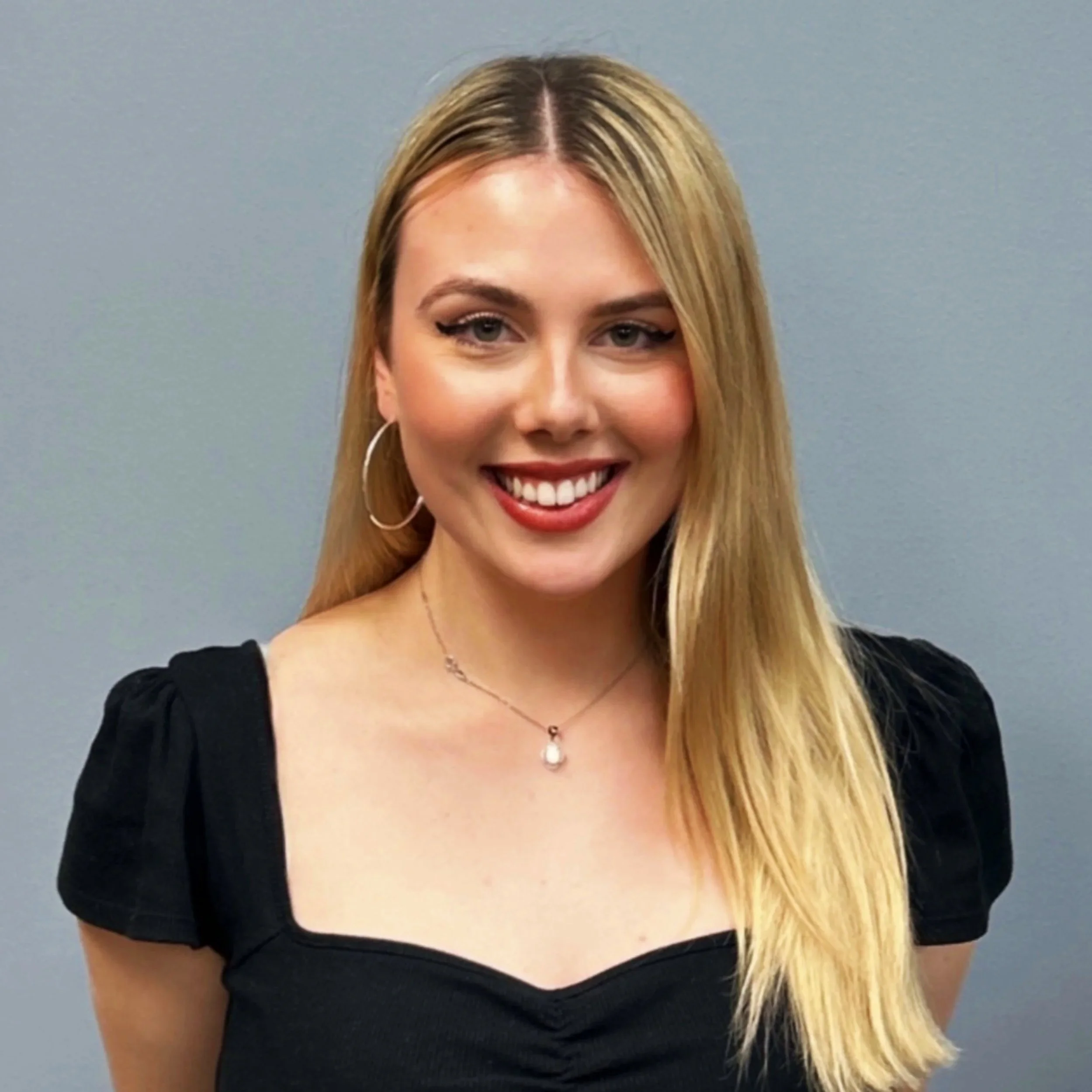 A woman with long blonde hair smiling and wearing makeup, a black top, a silver necklace, hoop earrings, and a white beaded necklace against a gray background.