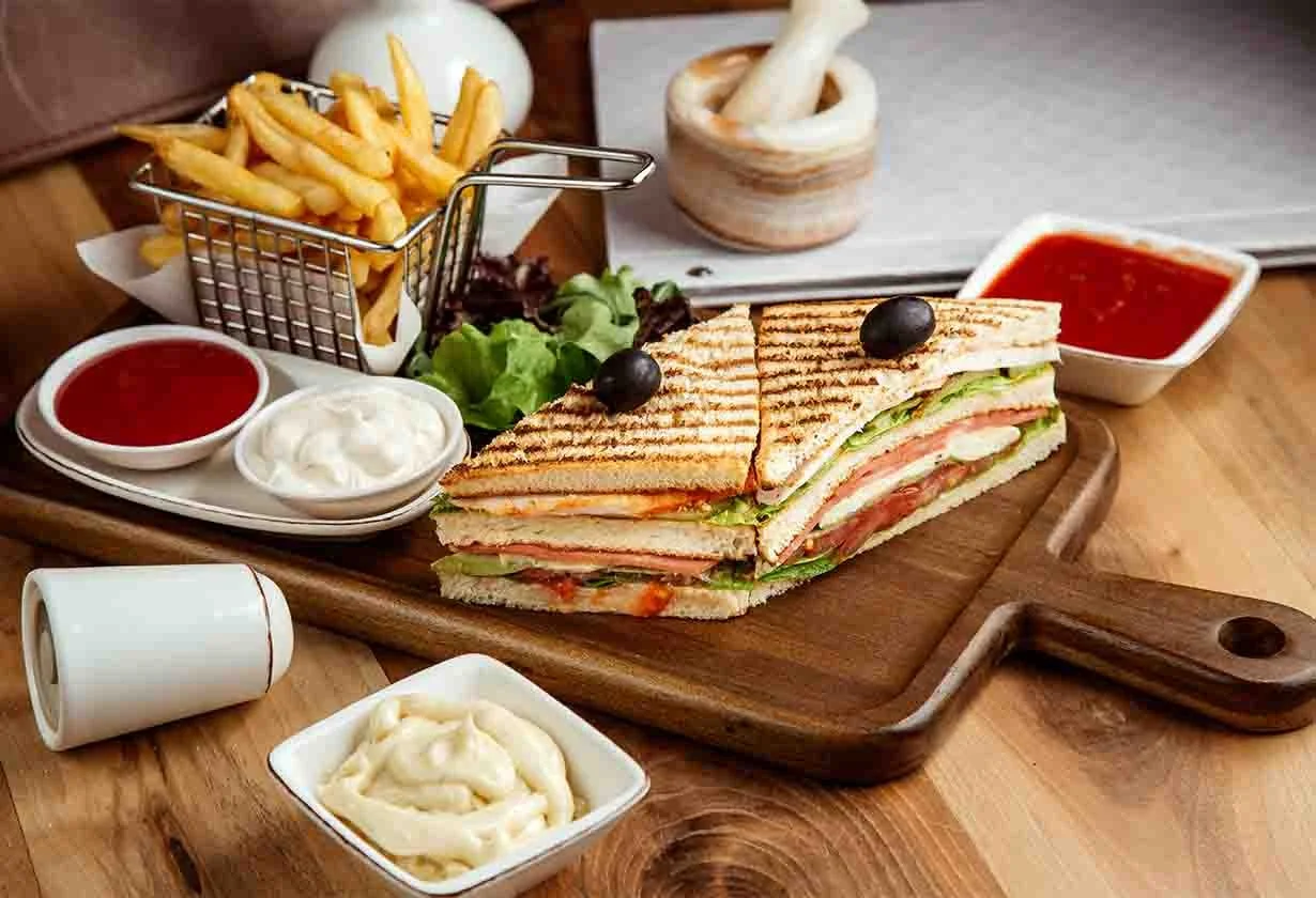 A club sandwich with grilled bread, lettuce, tomato, turkey, and cheese, garnished with olives, served on a wooden tray with multiple dipping sauces, served with fries and onion rings.