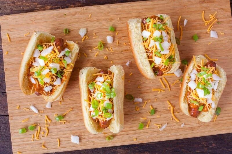 Four hot dog buns with chili, shredded cheese, chopped onions, and green onions on a wooden cutting board.