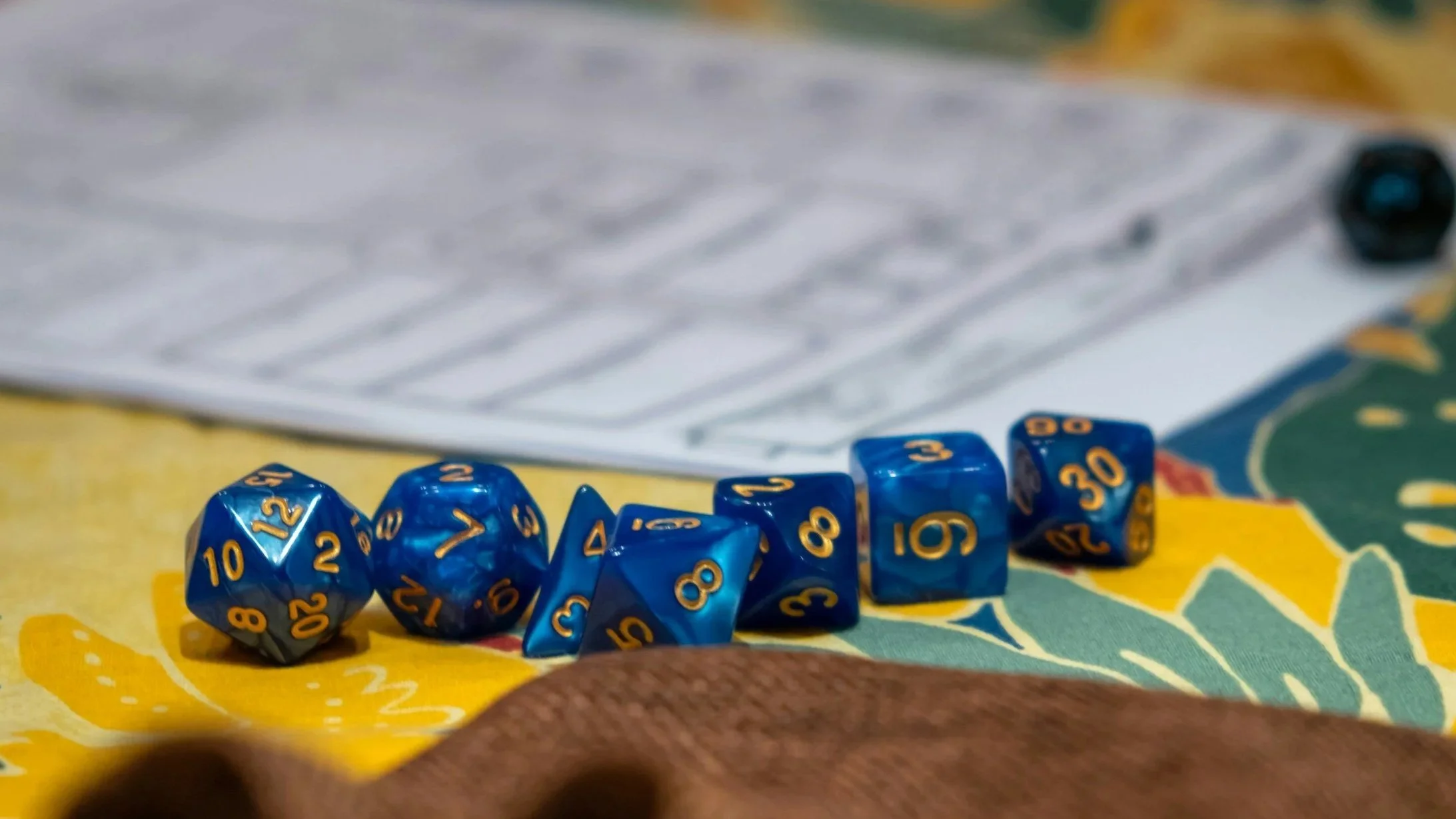 A collection of blue polyhedral dice with yellow numbers, including d10, d20, d6, d8, and d12, is arranged on a colorful tablecloth with a paper in the background.