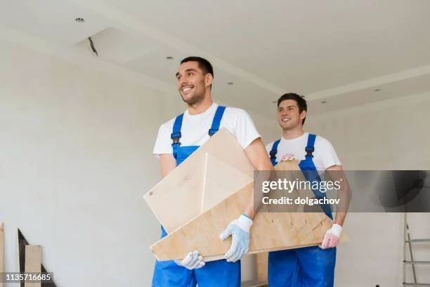 Two young men in white shirts and blue overalls carrying a large piece of marble or stone indoors.