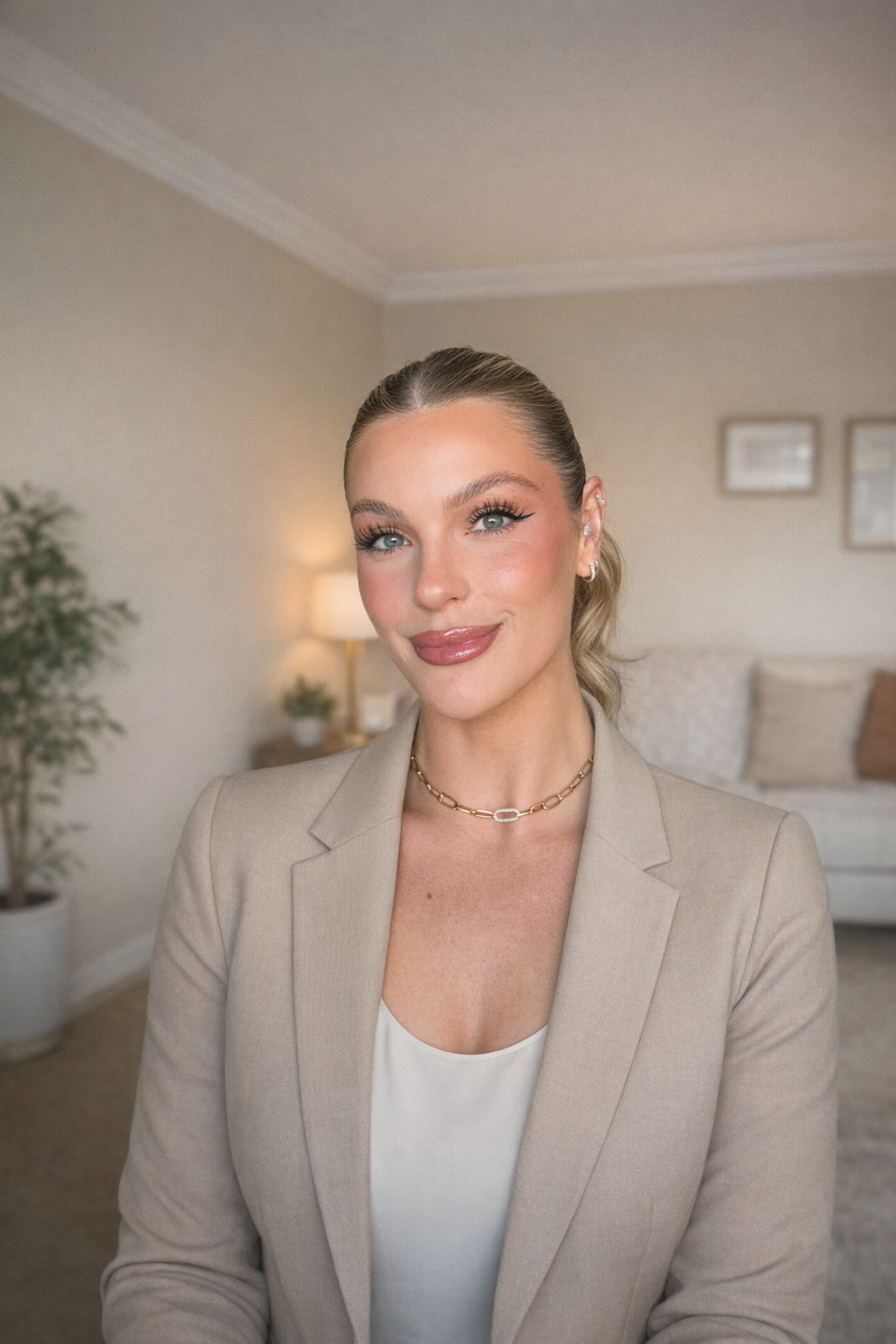A woman with blonde hair, blue eyes, and makeup, wearing a beige blazer, a white top, and a gold necklace, smiling in a living room with neutral-colored walls, a beige couch, and framed pictures.