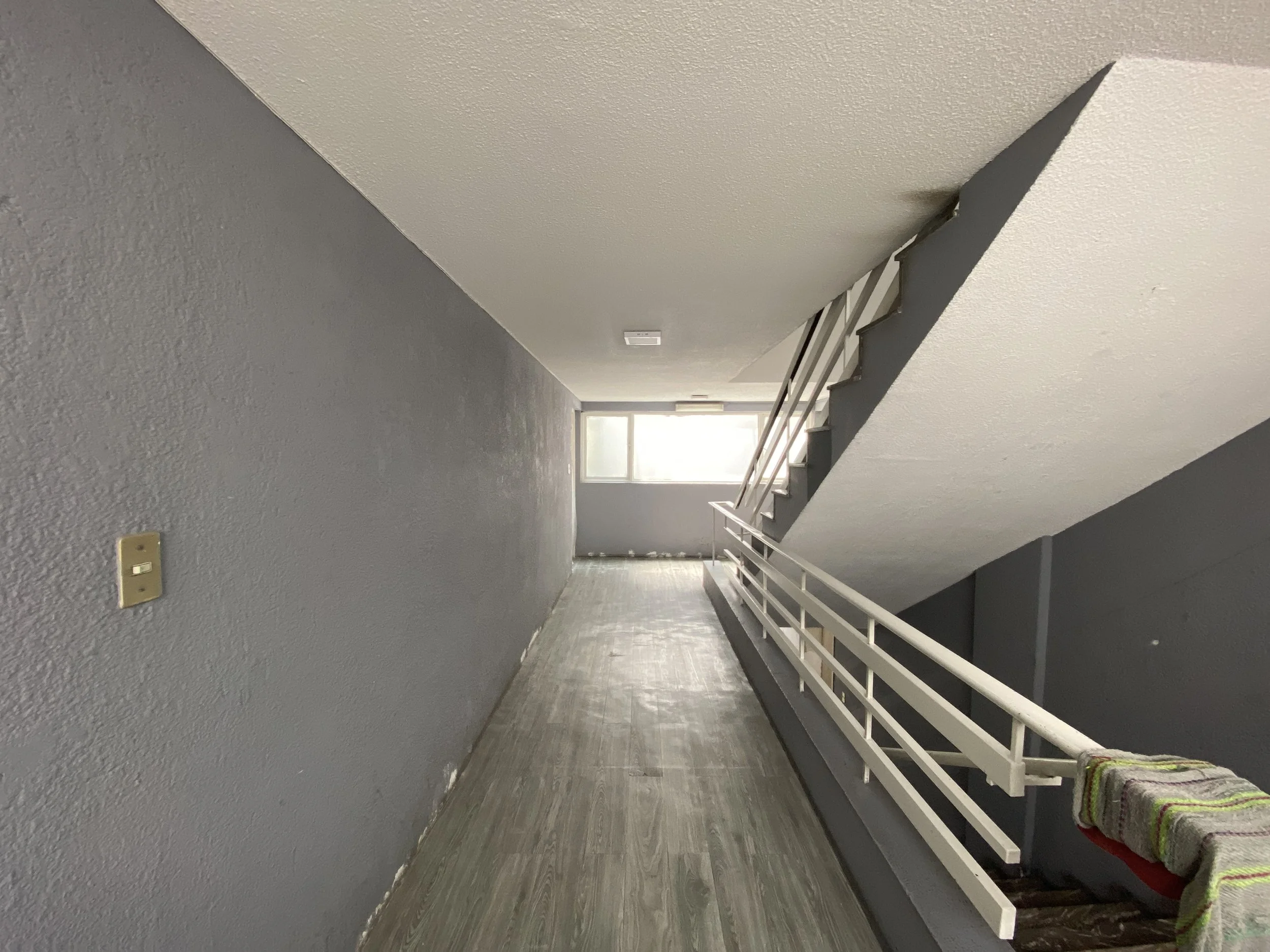 A long corridor with gray textured walls, a wood-patterned floor, a white staircase with towels on the bottom step, and a large window at the end letting in sunlight.
