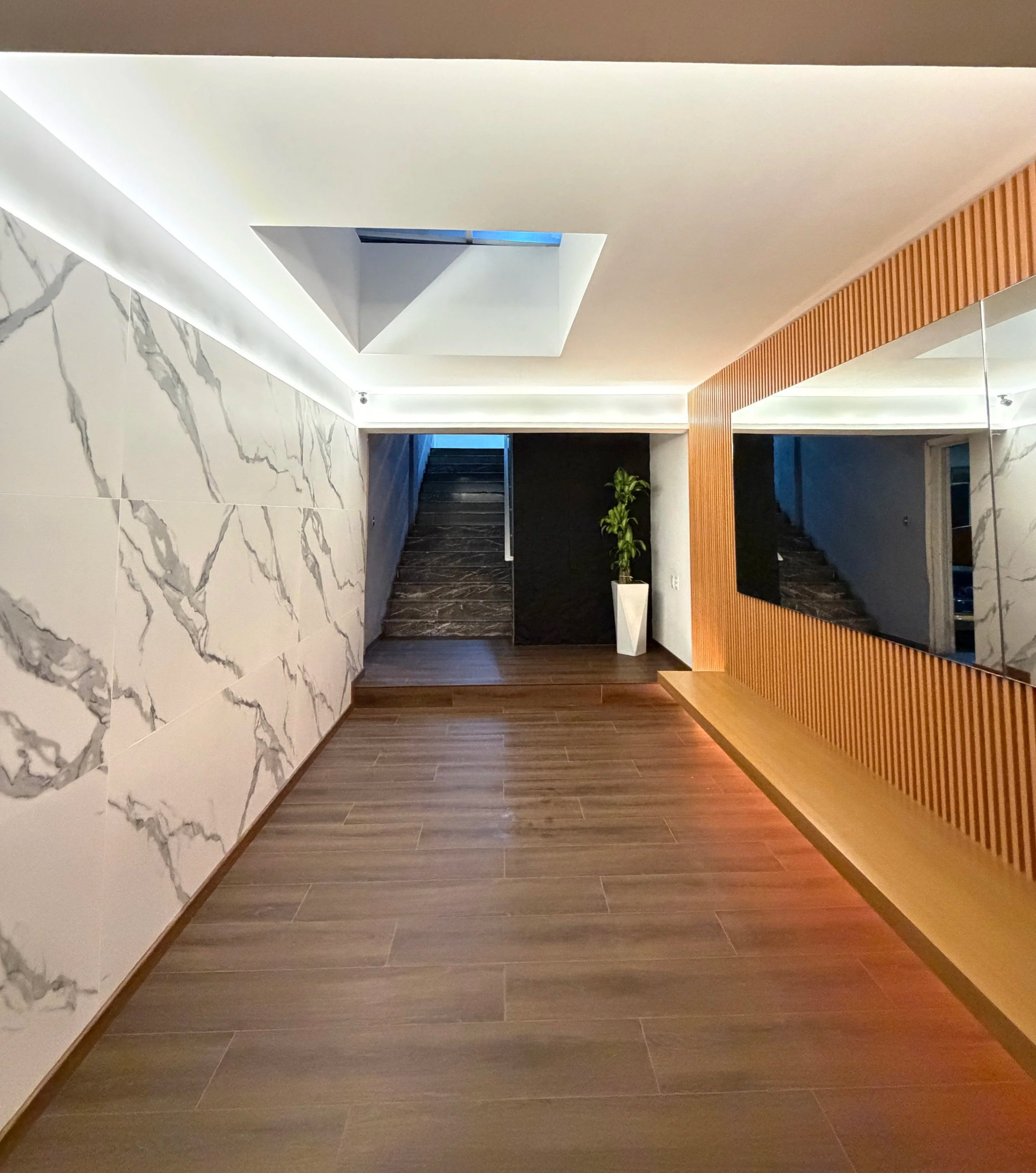 Modern interior lobby with white marble wall, dark wood flooring, and a wooden slatted wall feature. A potted plant is visible near stairway in the background.