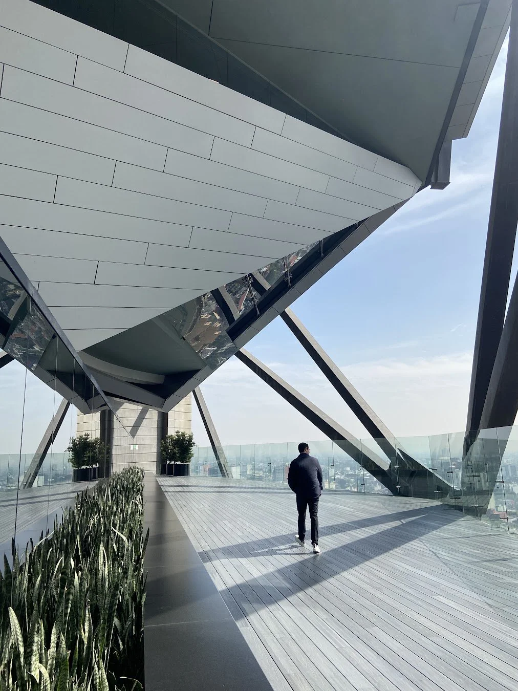 A person walking on a spacious outdoor terrace with modern architectural design, glass barriers, and a view of a city skyline in the background, overcast sky, and contemporary building elements.