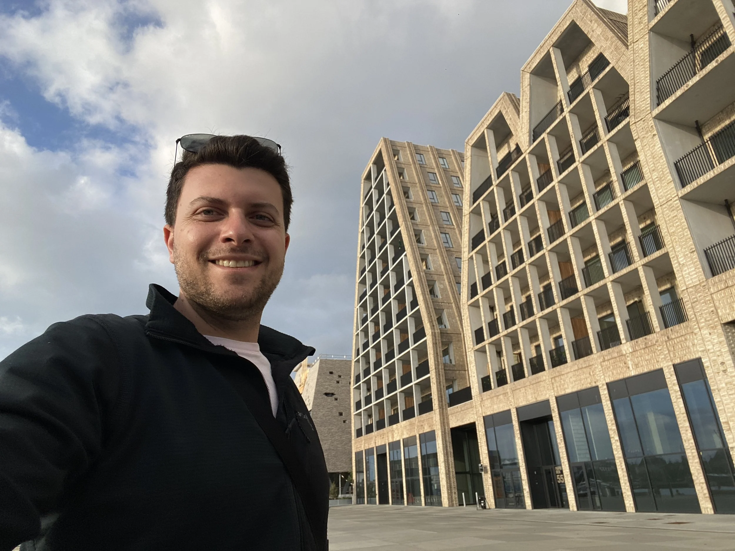 A man with short dark hair and sunglasses on his head is smiling in front of modern multi-story apartment buildings with large glass windows and balconies, under a partly cloudy sky.
