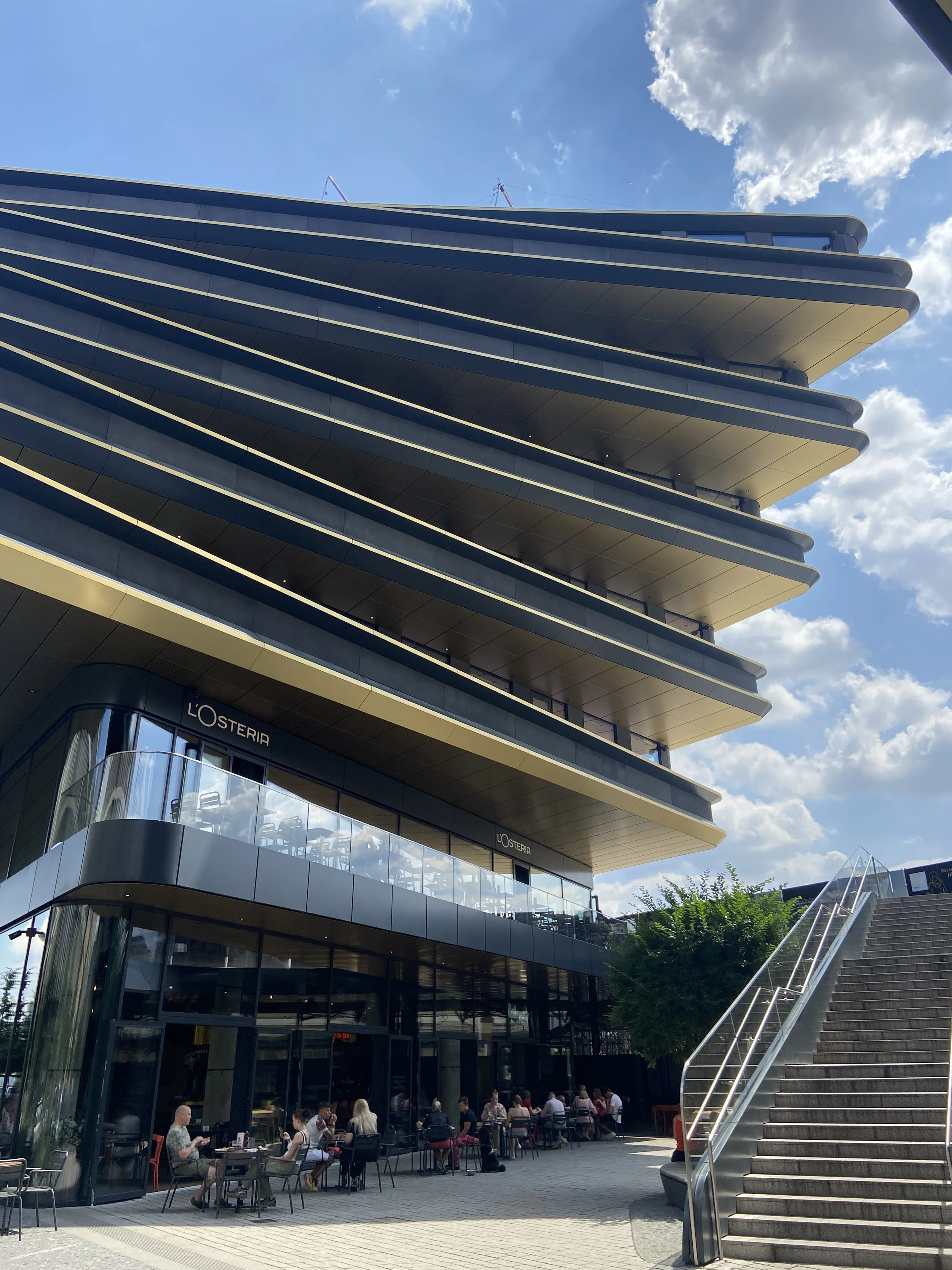 Modern multi-story building with outdoor seating area filled with people, a staircase to the right, and blue sky with clouds overhead.
