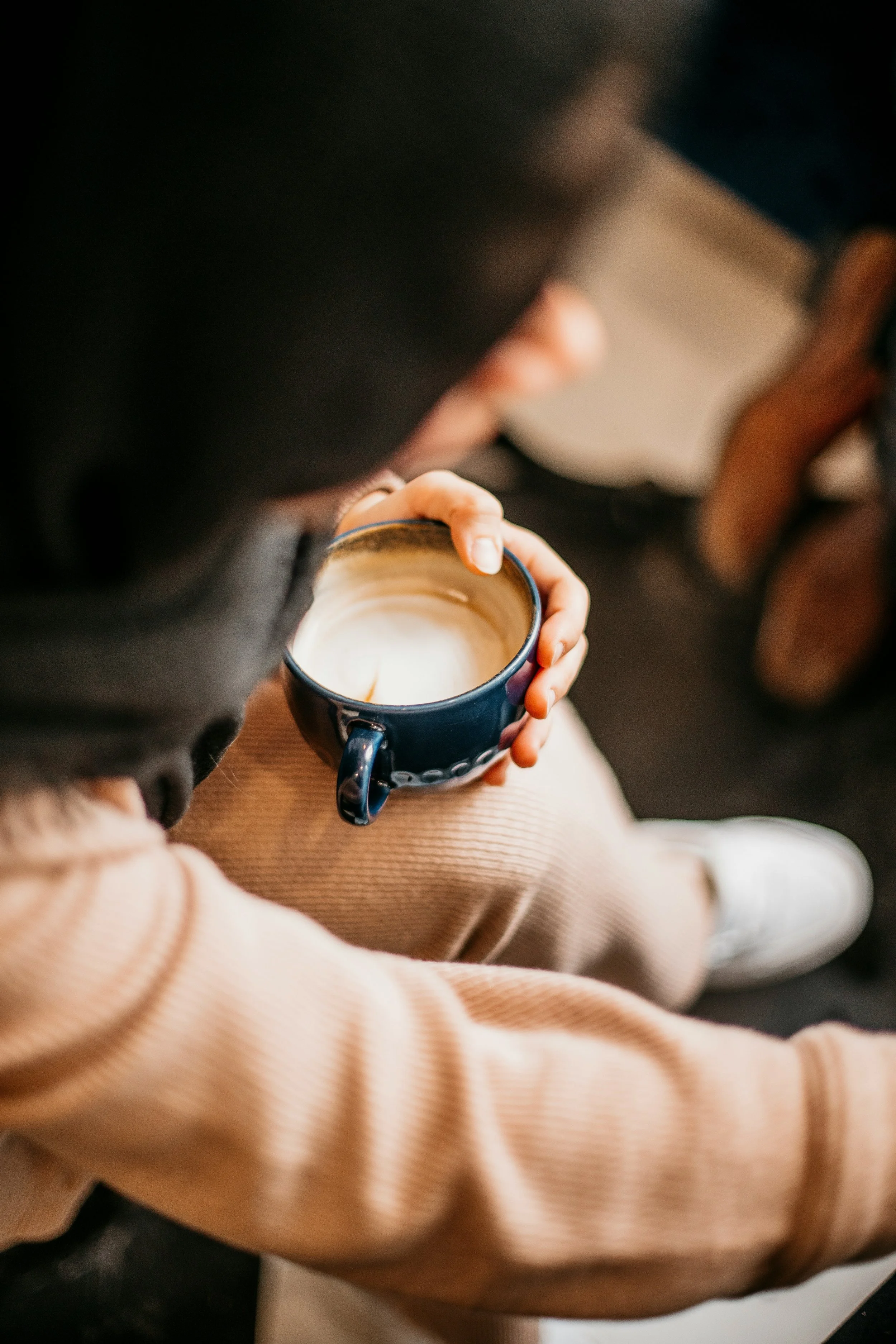 Person wearing beige pants and a beige sweater, holding a ceramic cup with a dark exterior and cream-colored interior, with a beverage inside, sitting on a dark surface.