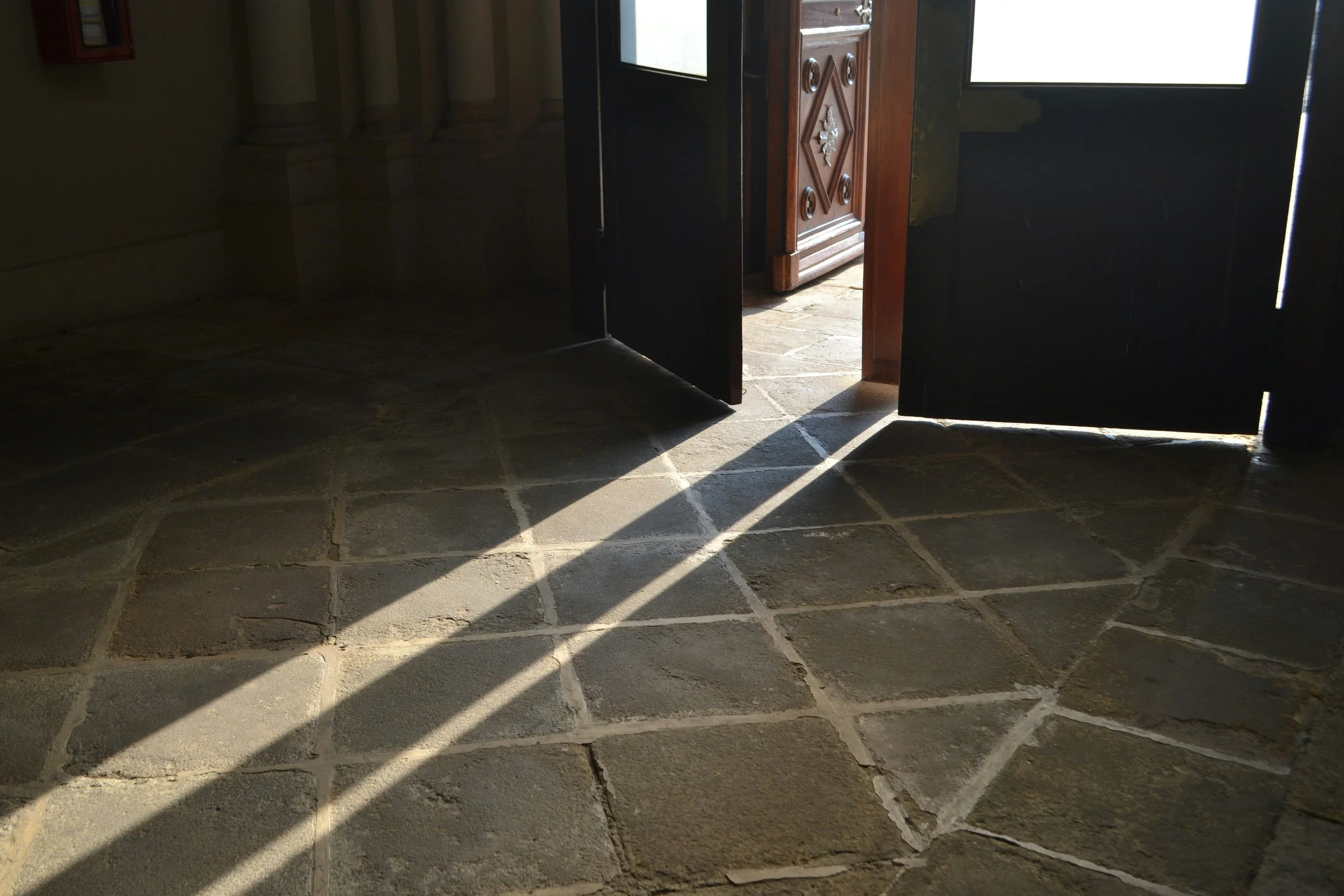 An arched stone entryway with one door swung open, sunlight cutting diagonally across the flagstone floor toward a carved wooden inner door visible through the opening.