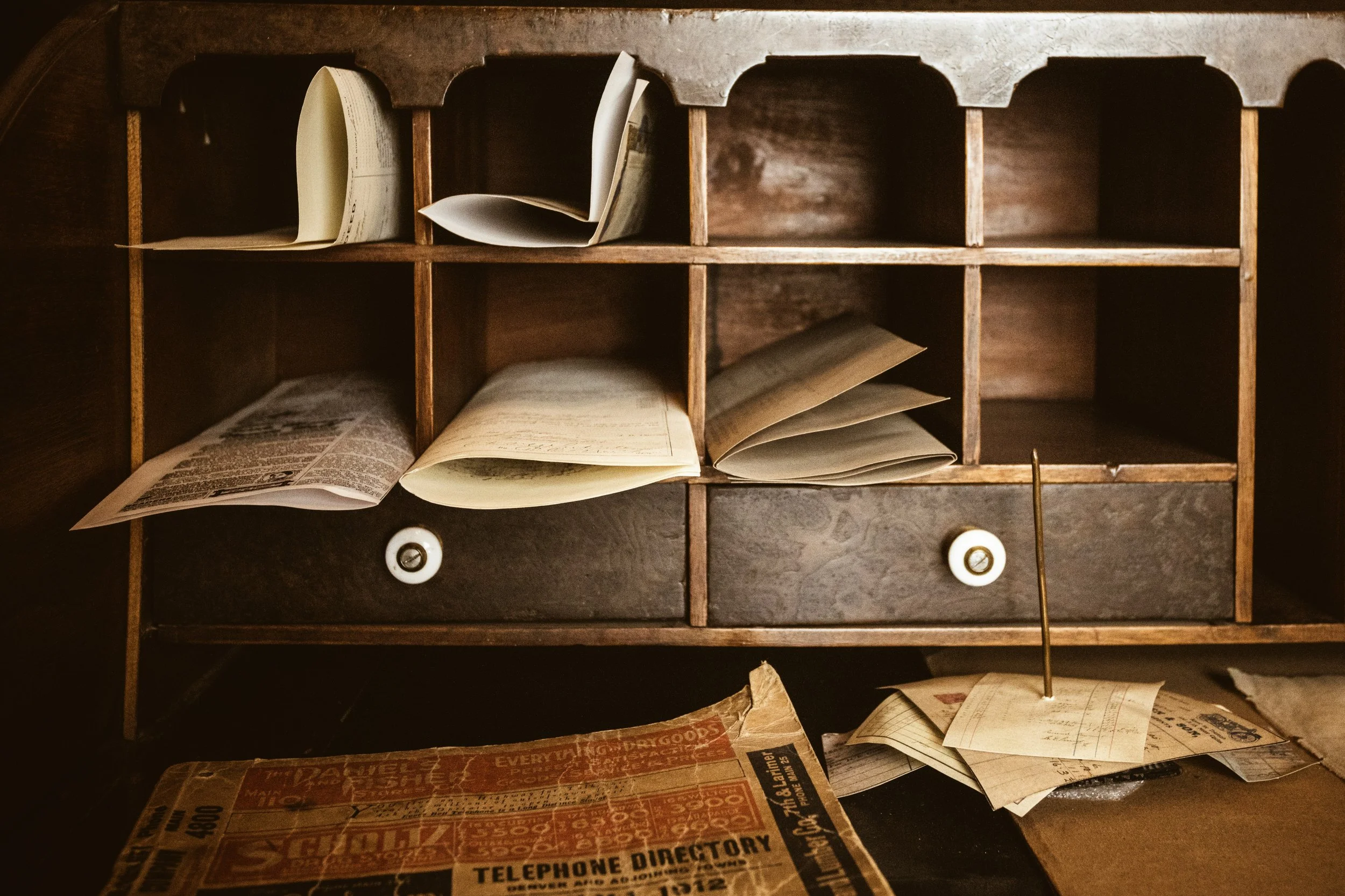 A worn wooden desk organizer with papers tucked into pigeonhole slots, a pair of small drawers, and an old telephone directory on the surface below.