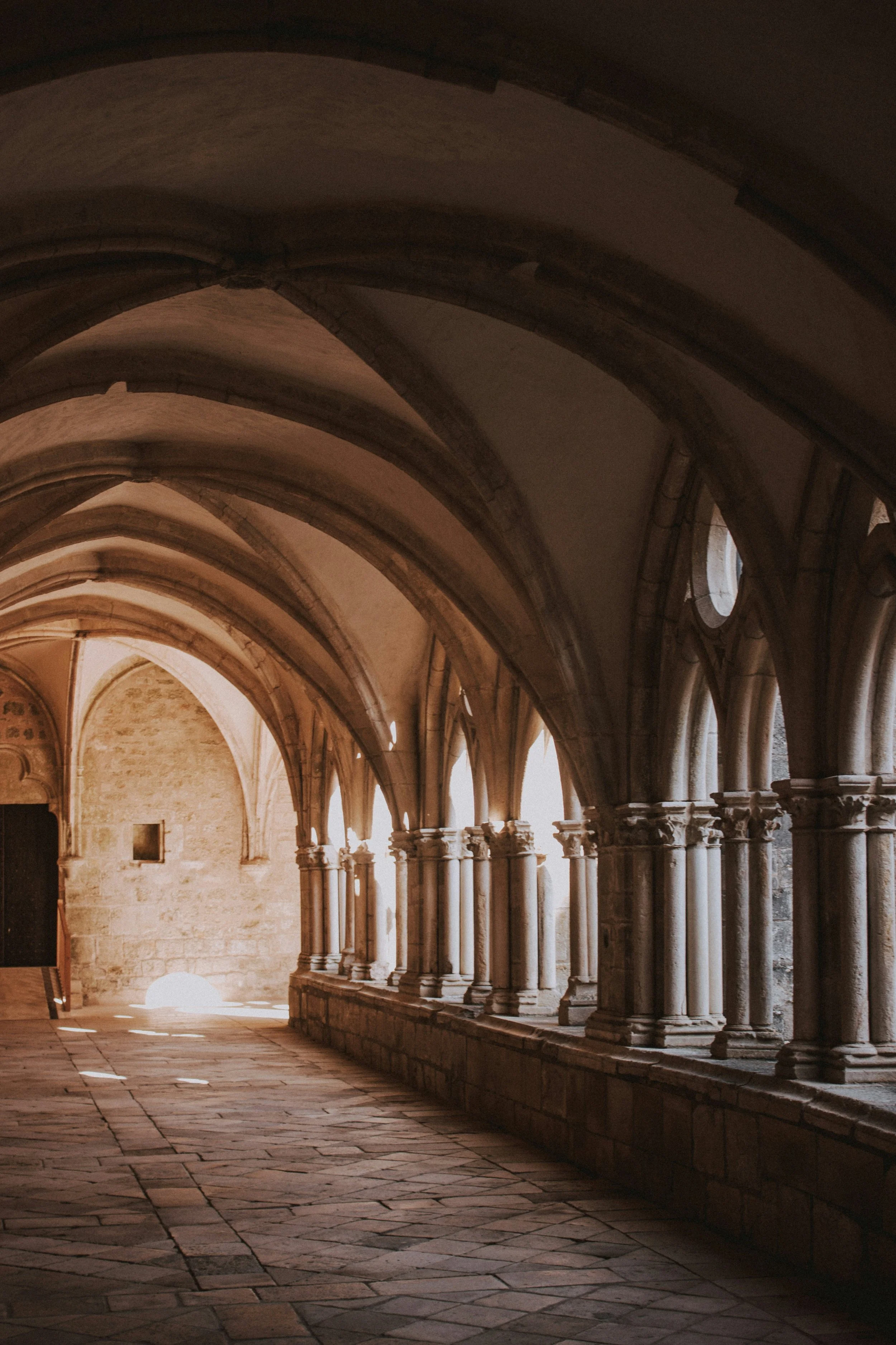 The interior of a historic stone cloister, with repeating vaulted arches overhead and soft afternoon light falling across the flagstone floor.