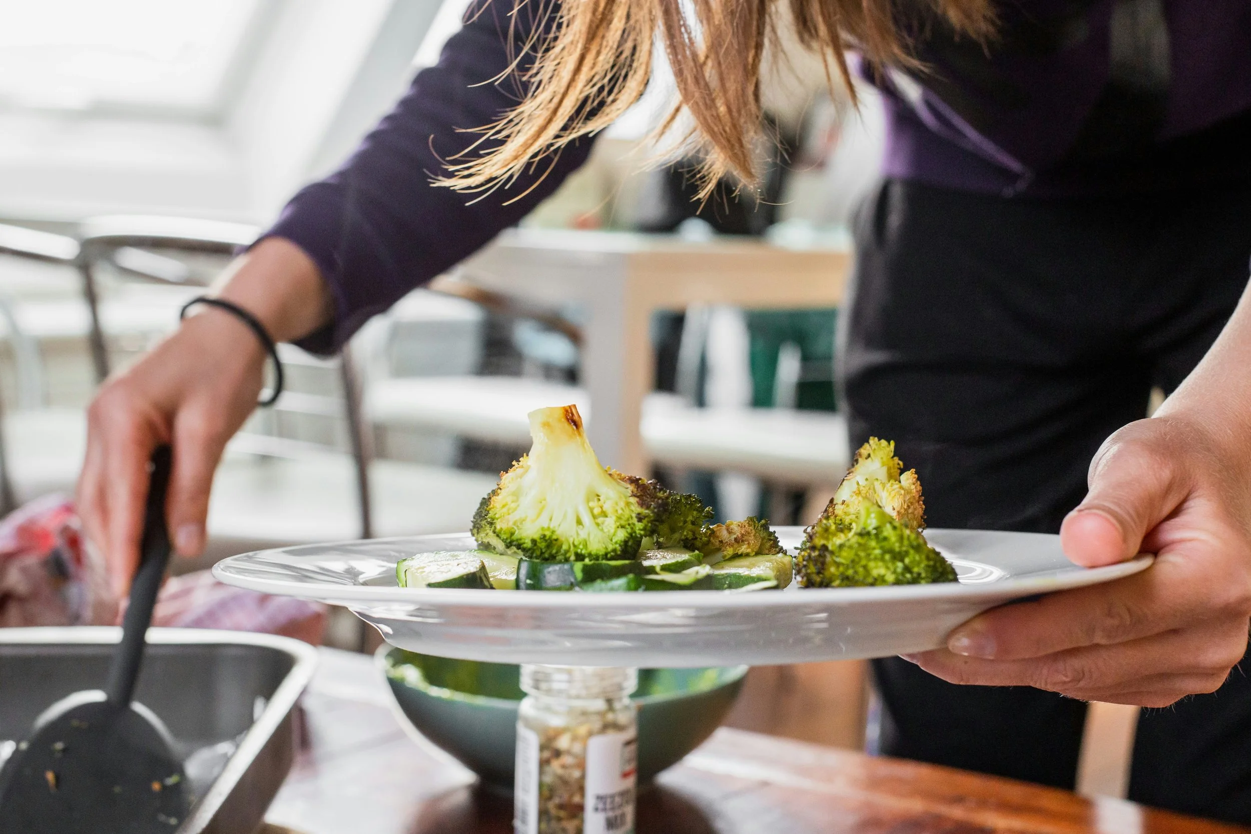 Person serving broccoli and cucumbers on a plate in a kitchen setting.